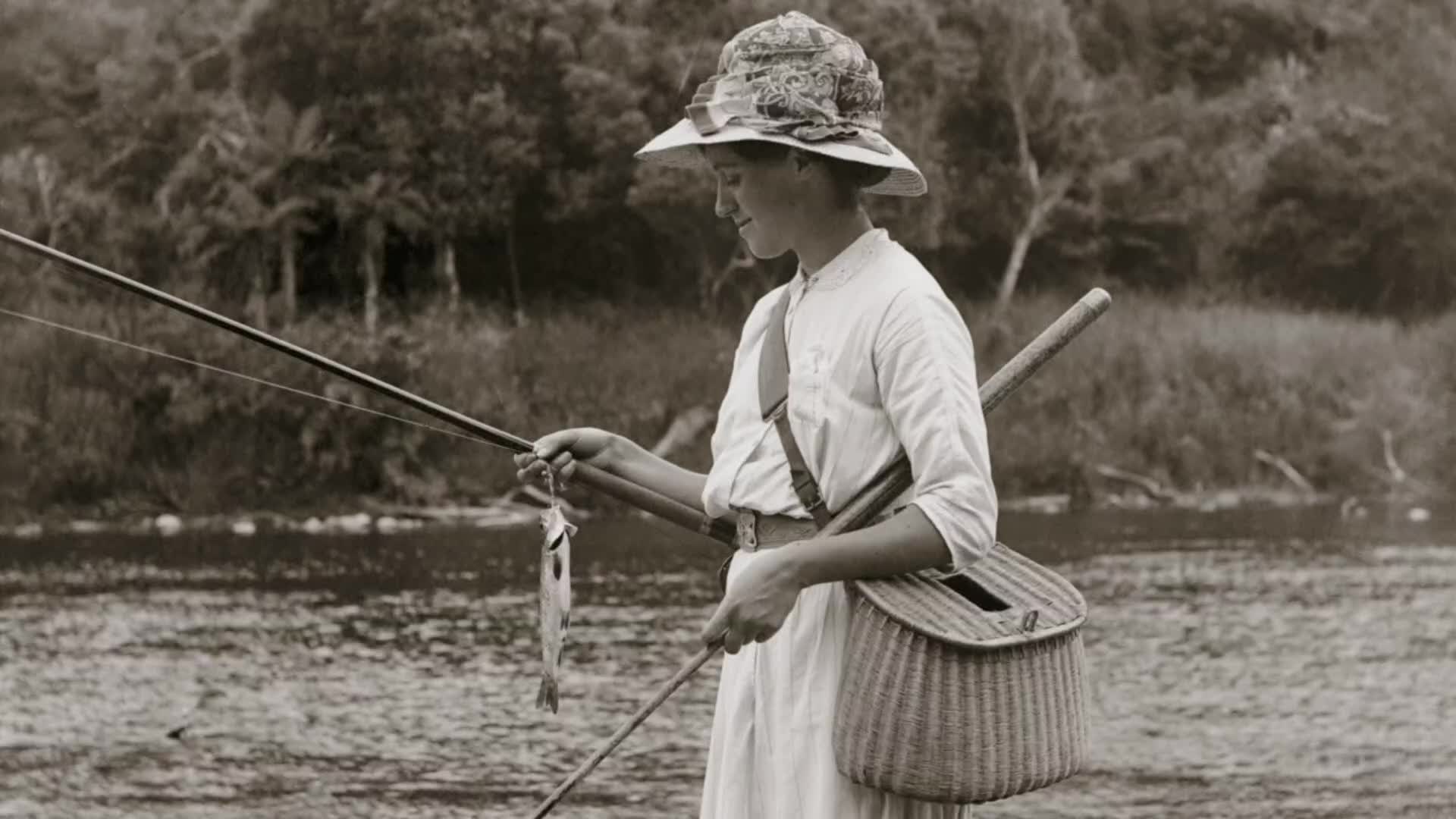 A young woman, dressed for a day in the New Zealand countryside, holds a small fish on her line. She wears a wide-brimmed hat and a basket hangs from her shoulder. A young woman, dressed for a day in the New Zealand countryside, holds a small fish on her line. She wears a wide-brimmed hat and a basket hangs from her shoulder.