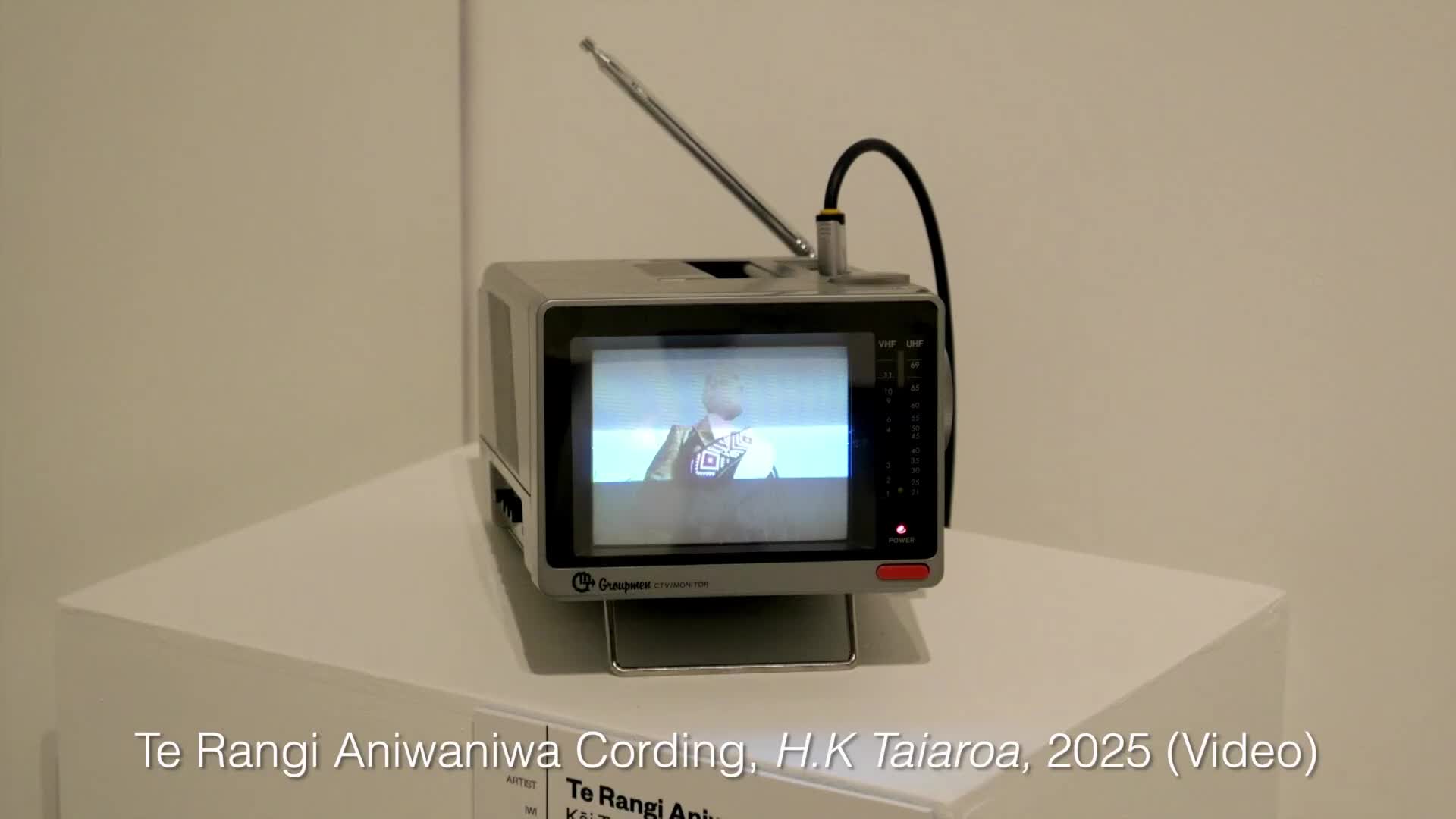 A small, grey television sits on a white pedestal, its screen showing a person in what looks like a parliamentary setting. The antenna is extended, and a black cable connects to the top of the set. A small, grey television sits on a white pedestal, its screen showing a person in what looks like a parliamentary setting. The antenna is extended, and a black cable connects to the top of the set.