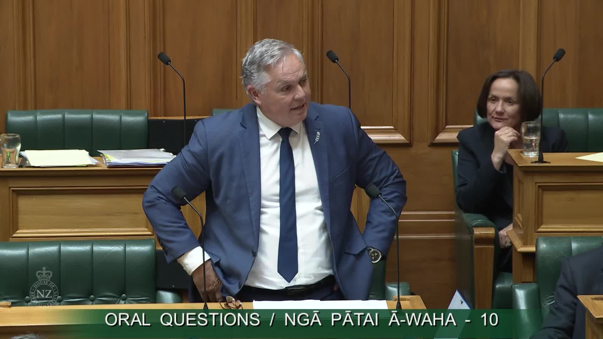 A man in a blue suit with a dark tie speaks, his hands resting on his hips. Beside him, a woman in a dark jacket watches with a slight smile, her chin propped on her hand.