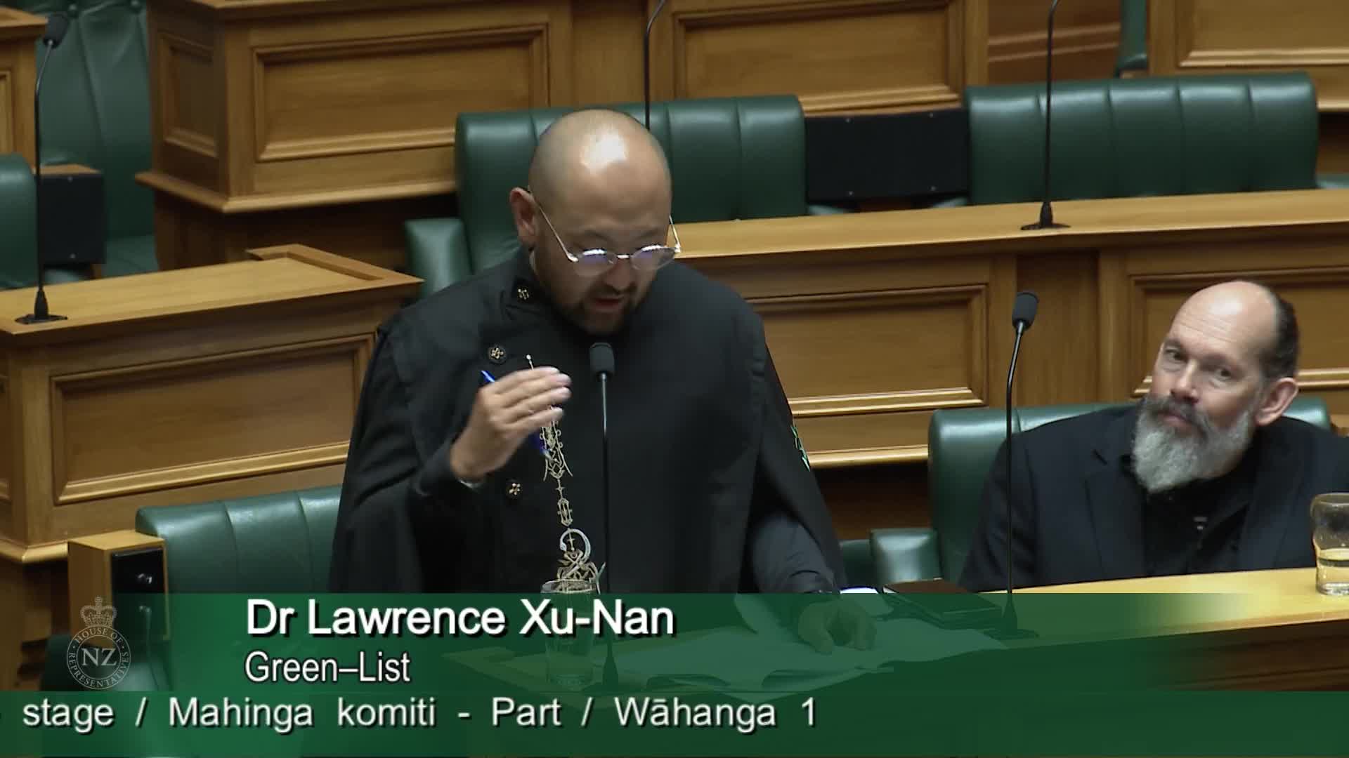 Dr Lawrence Xu-Nan, a Green-List MP, speaks into a microphone. A man with a grey beard watches him from the side.