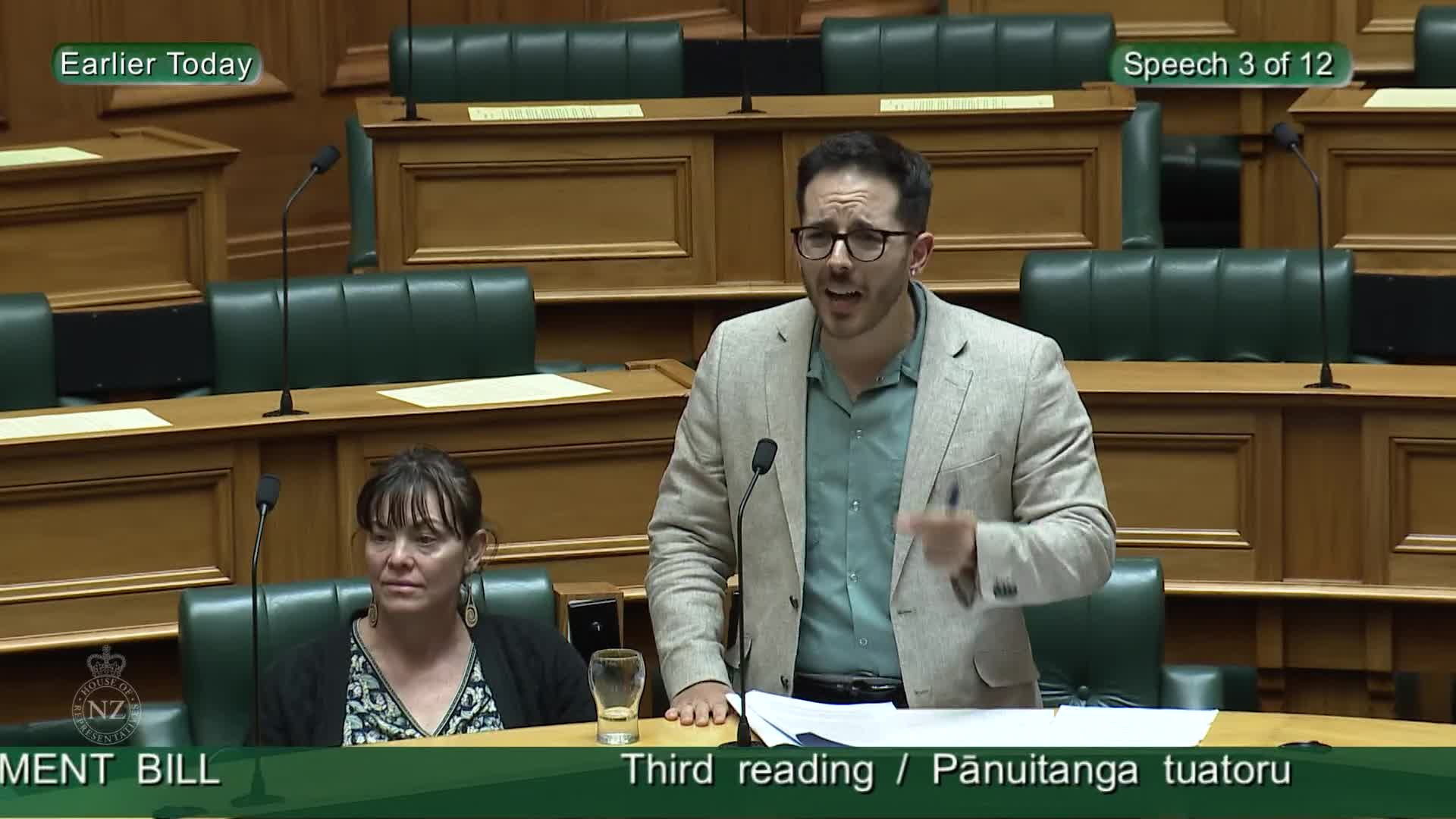 A man in a light jacket stands, gesturing with his right hand as he speaks into a microphone. Beside him, a woman sits, looking forward with a neutral expression. This is the Parliament of New Zealand, broadcasting live.