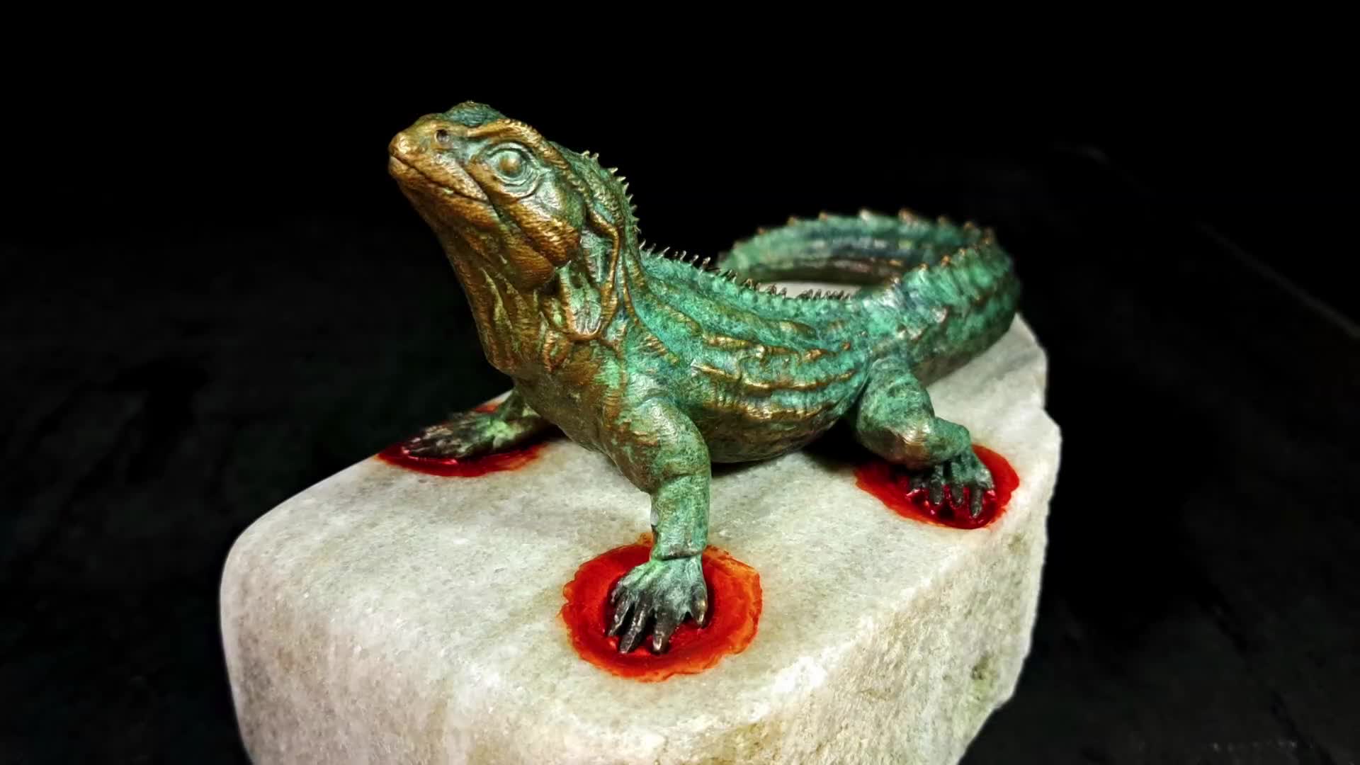 A bronze tuatara, its scales a verdigris green, rests on a rough, pale stone. Red droplets cling to its feet, stark against the rock.