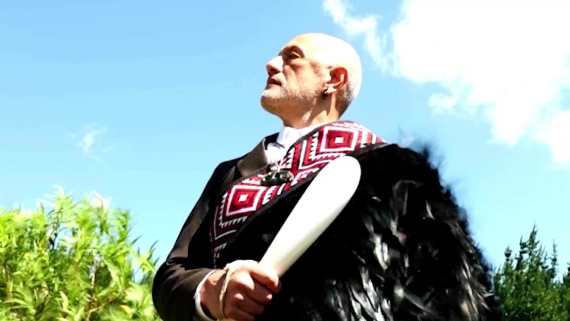 A man stands with a carved wooden club, a korowai draped over his shoulders. He looks up at the bright New Zealand sky, a slight smile on his face.