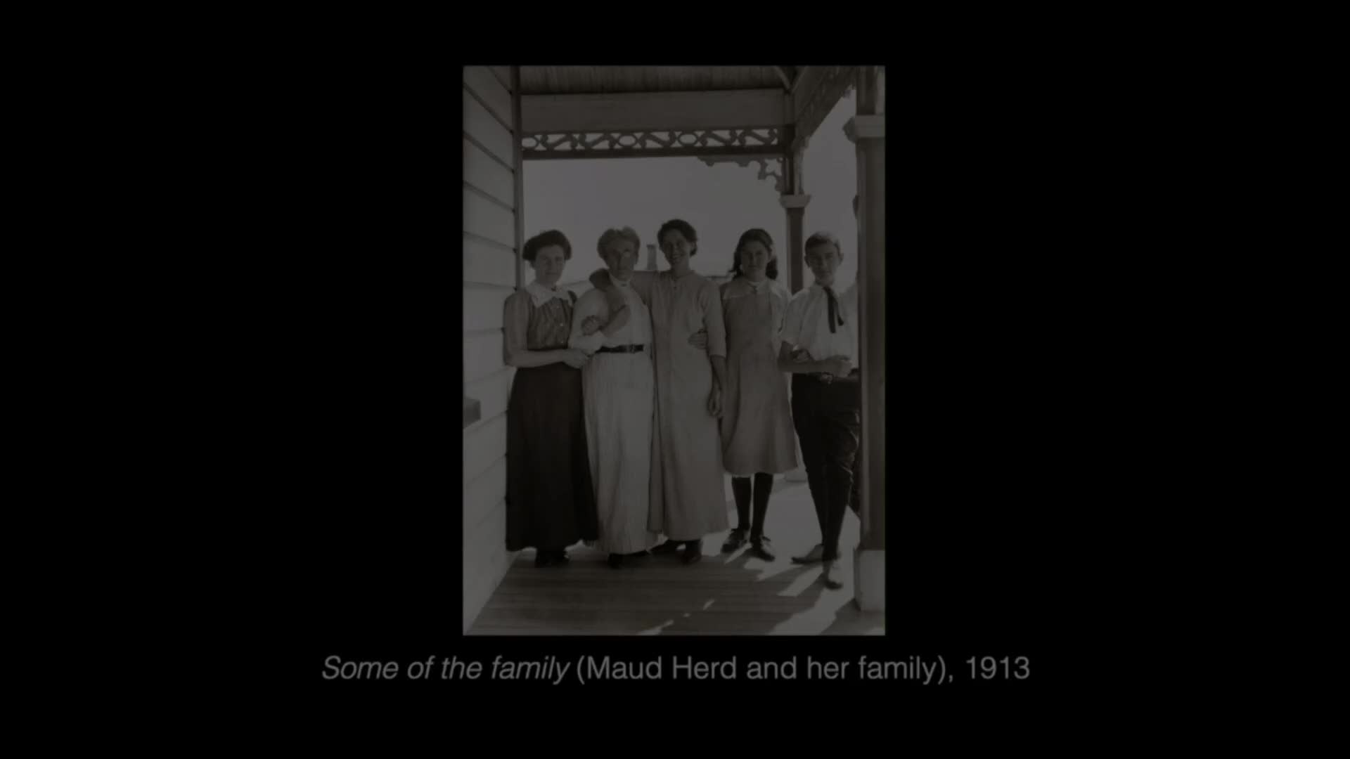Five people stand on a porch, dressed in early 20th-century New Zealand fashion. The women wear long skirts and high-necked blouses, while the young man on the right sports a shirt with a tie.