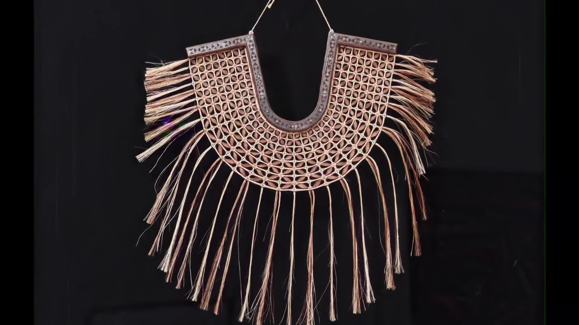 A woven flax collar hangs against a dark background. The intricate pattern of the collar, with its woven circles and dangling strands, is clearly visible.