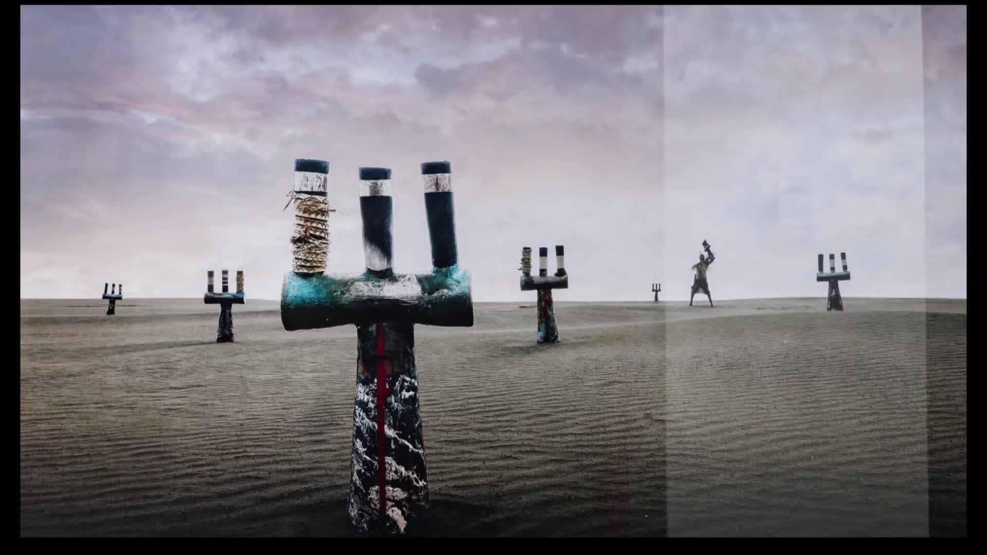 A lone figure stands in the distance, silhouetted against the grey sky. Around them, weathered wooden structures rise from the sand, their forms stark against the desolate landscape.