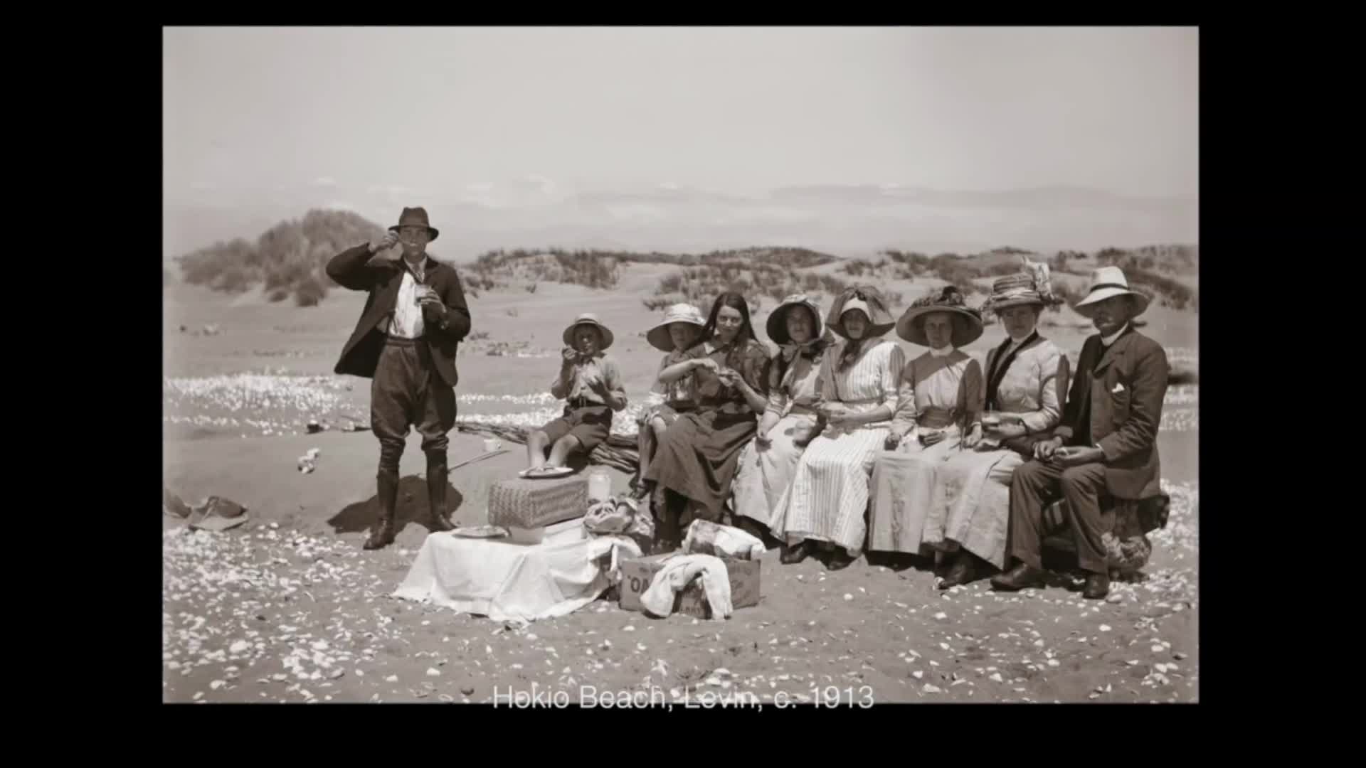 A man in a hat and jacket stands on the beach, lifting something to his lips. A group of women and children sit together on the sand, their long dresses and hats a clear sign of the era.