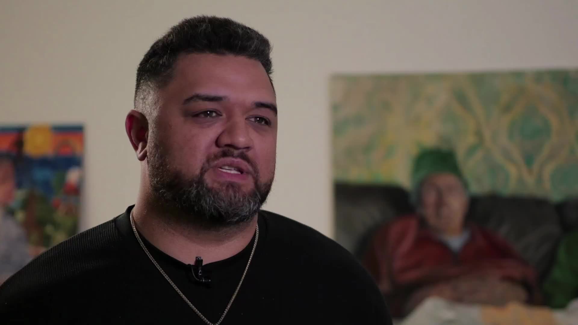A man with a beard speaks directly to the camera, his black shirt and silver chain visible. Behind him, two paintings hang on the wall, one featuring abstract colours and the other a blurred portrait.