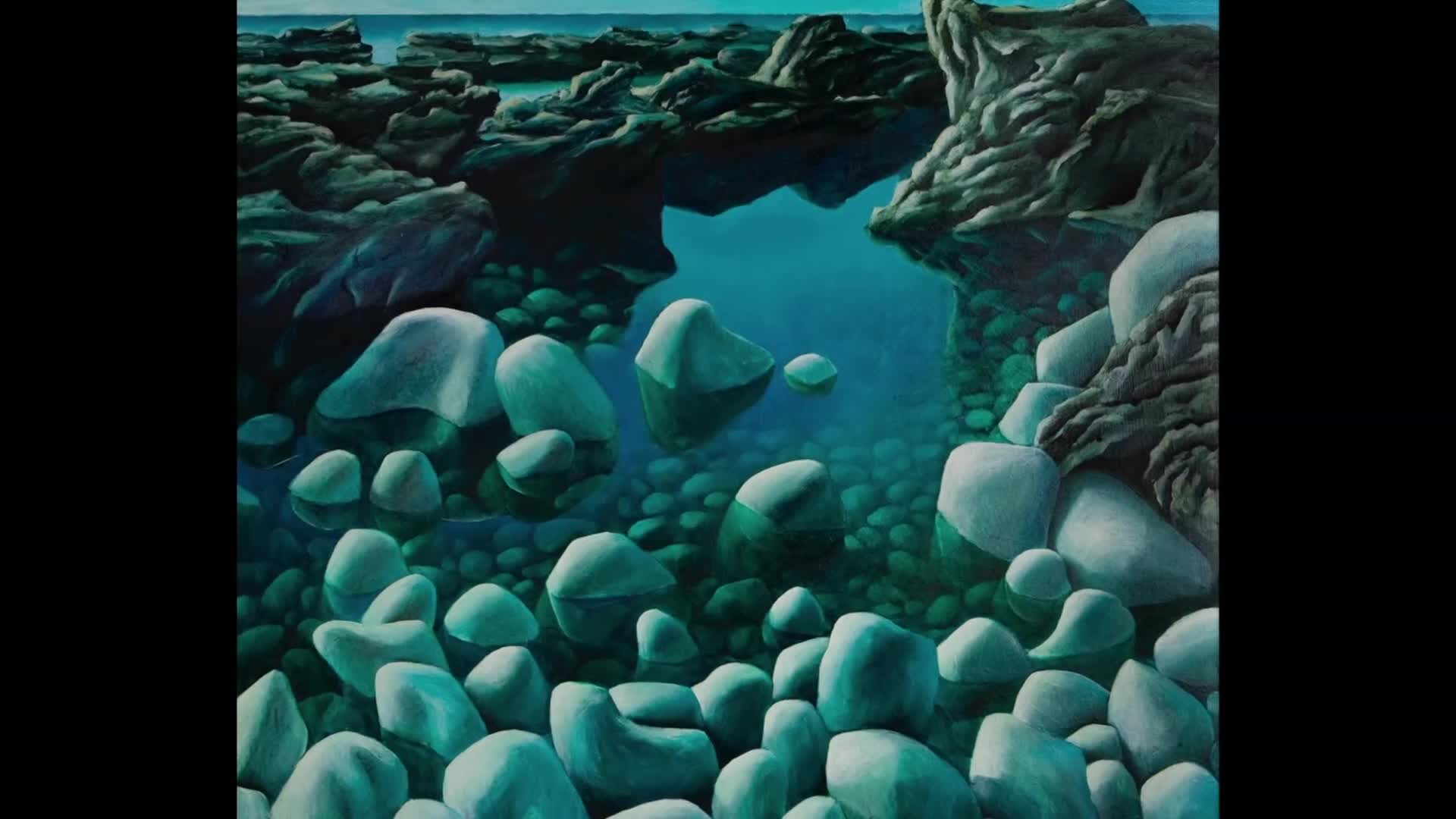 Smooth, grey rocks fill the foreground, submerged in clear, turquoise water. The water reflects the sky and the dark, rugged rocks that form the shoreline.