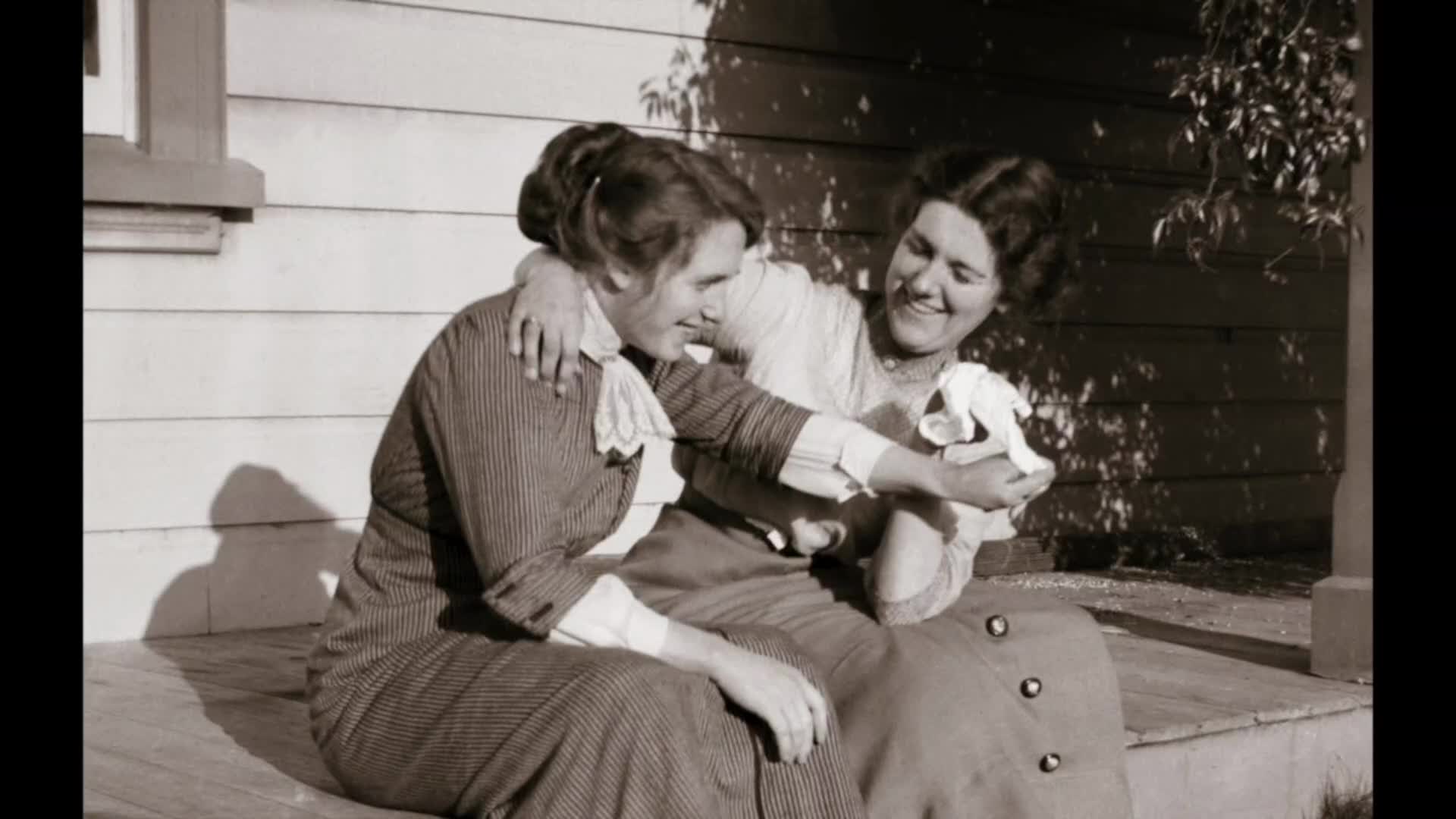 Two women sit on a porch, one with her arm around the other's shoulders. The woman on the right laughs, holding a small white bundle.