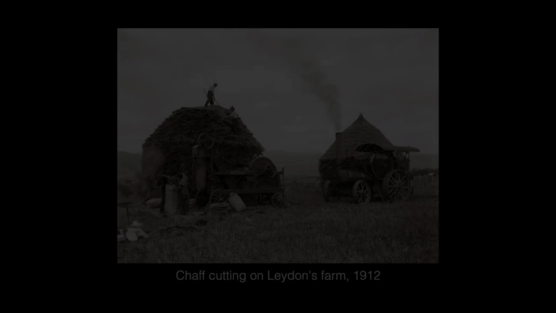 A man stands atop a massive stack of chaff, while a steam engine nearby belches dark smoke into the overcast sky. This is Leydon's farm, 1912.
