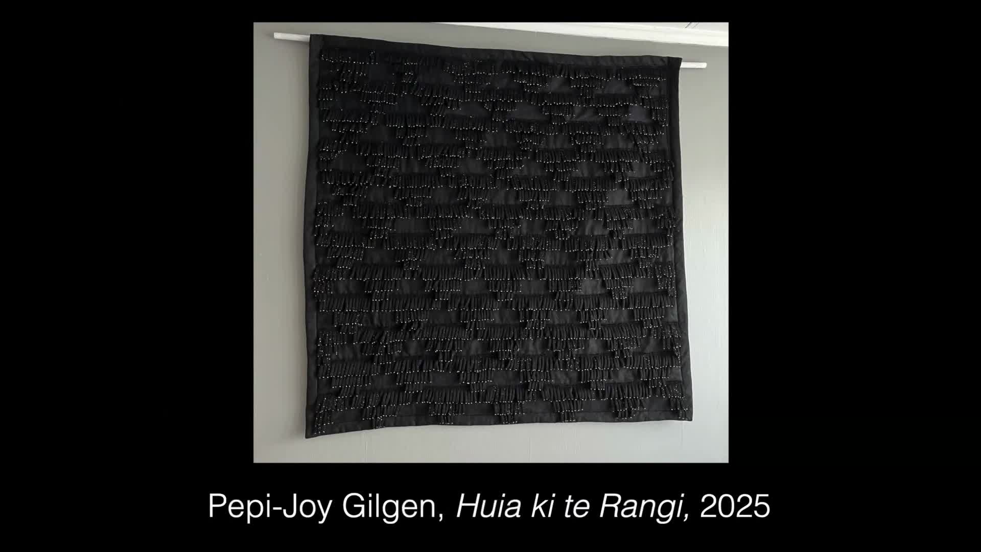 The black fabric hangs, its surface covered in rows of tiny, dark beads. Each bead is attached to a small fringe, creating a textured, almost shimmering effect across the artwork.