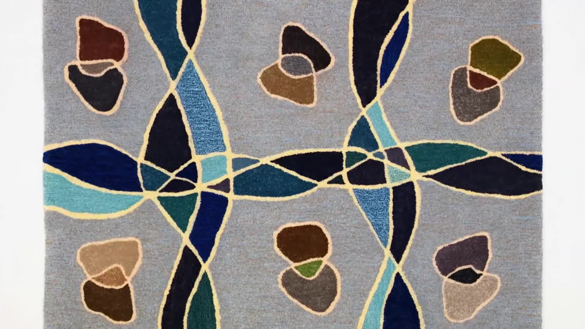 The woven tapestry hangs, a vibrant display of intersecting lines and abstract shapes.  Dark blues and greens weave through lighter blues and yellows, all set against a soft grey background.