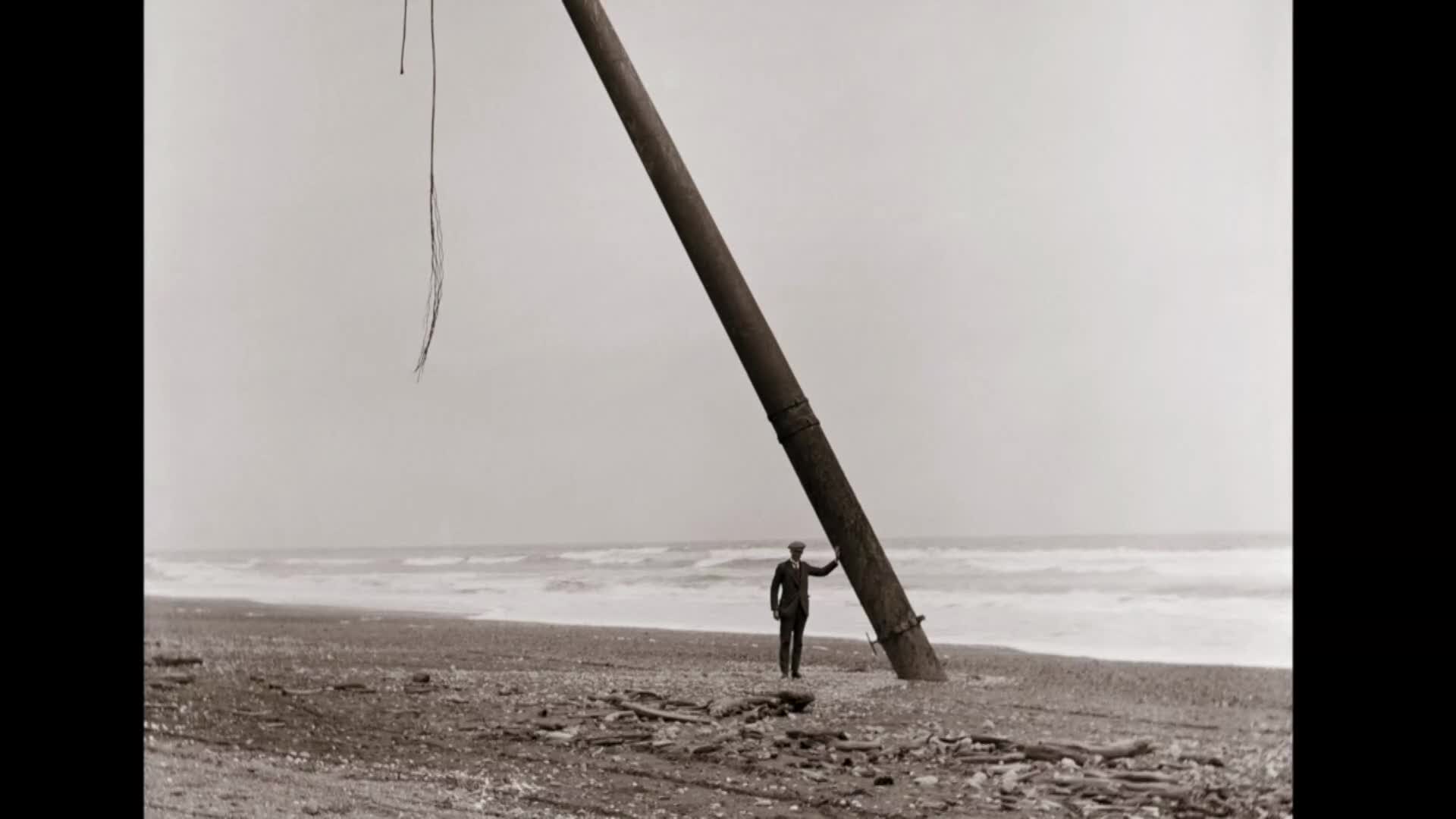 A man in a suit stands on a New Zealand beach, his hand resting on a thick, angled pole. Waves crash behind him, their white foam stark against the grey sea.