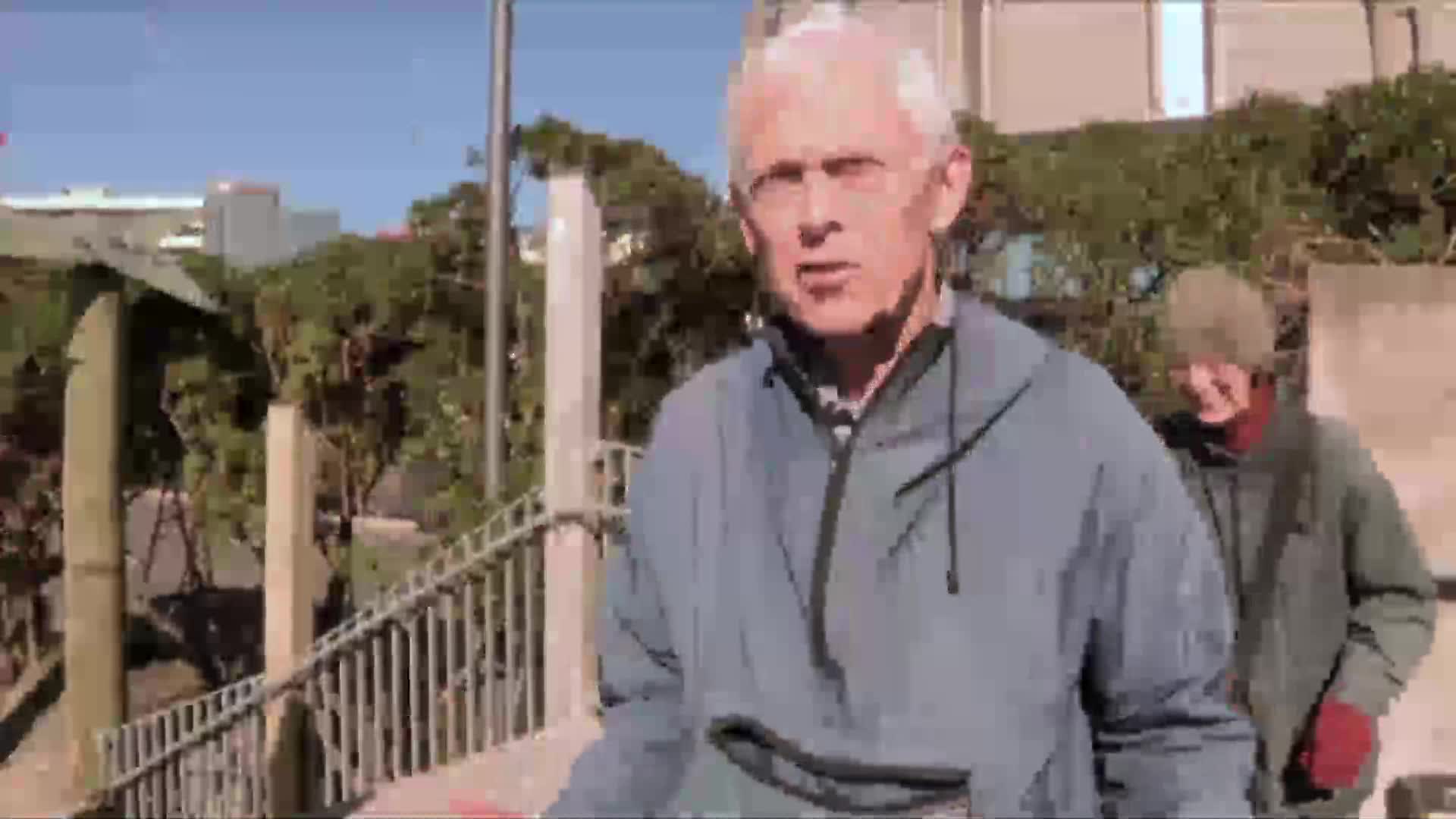 A man with white hair in a blue jacket is speaking, the camera focused on him. Behind him, another person in a hat and jacket walks casually down some stairs.
