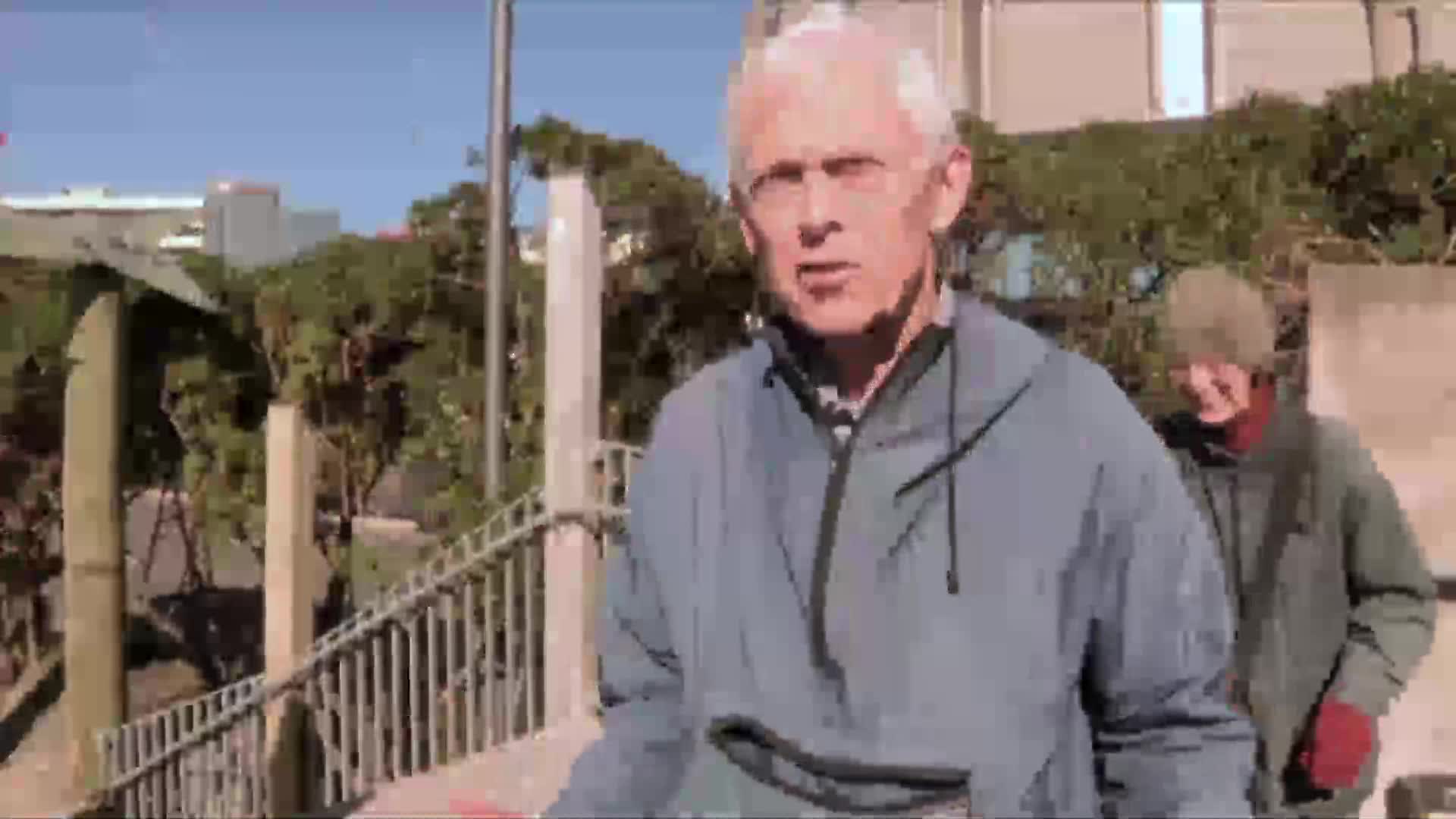 A man in a blue jacket is speaking, looking directly at the camera. Behind him, another person in a green jacket and a hat walks along a walkway.

