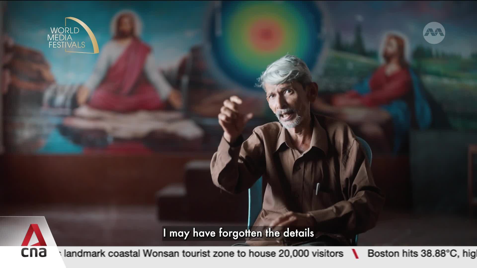 A man with gray hair and a brown shirt gestures with his hands as he speaks. Behind him, a mural of a religious figure is visible, and the CNA International logo is at the bottom.
