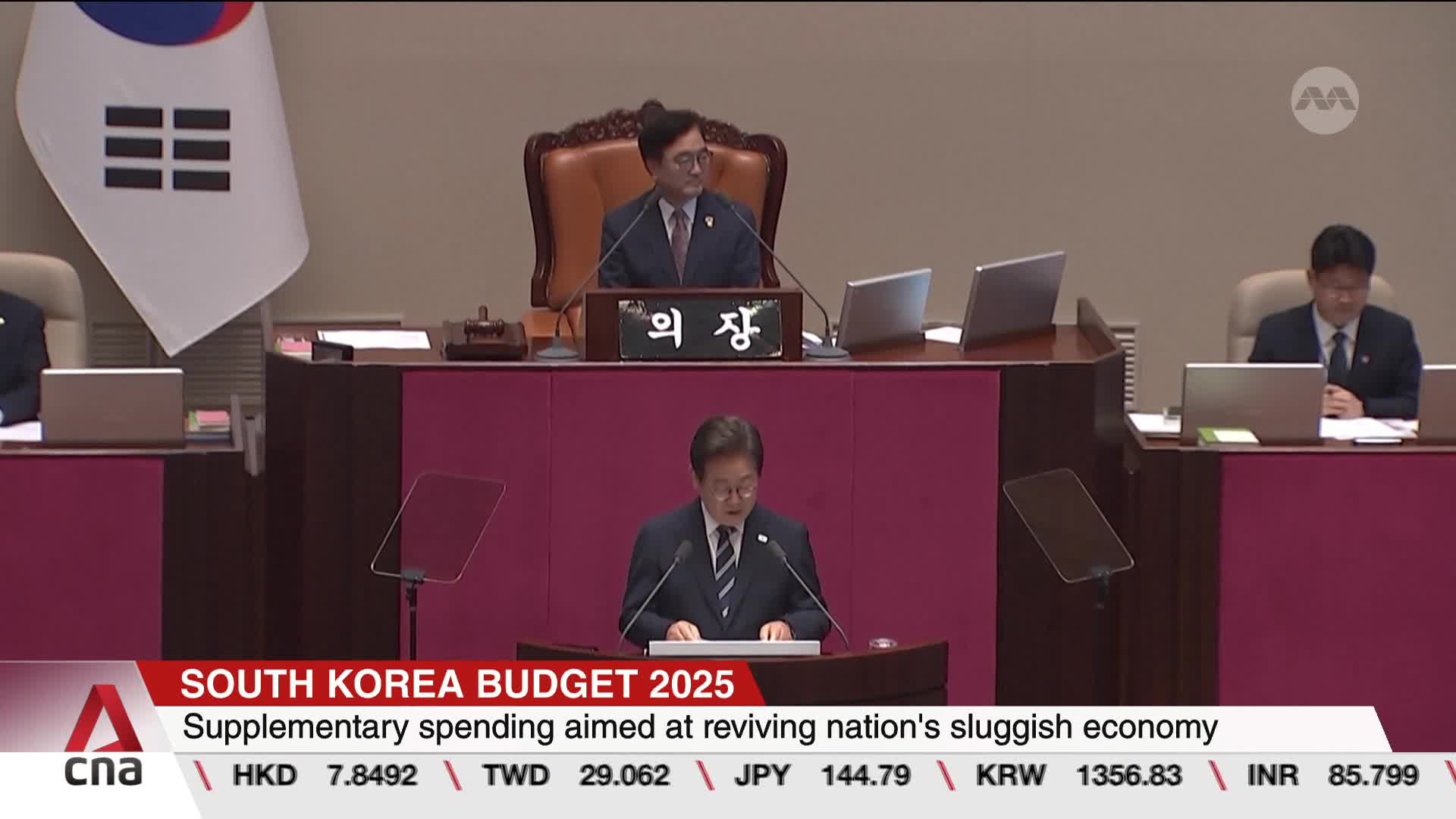 A man in a suit speaks from a podium, addressing the room.  Behind him, a flag with a black and white design hangs, and the CNA International graphic displays financial data.
