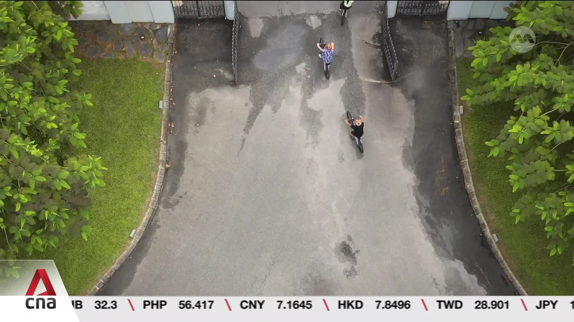From above, two people cycle along a wet driveway, heading toward a wrought-iron gate. A third person is further back, also on a bike, and the scene is framed by green foliage and the CNA International logo.
