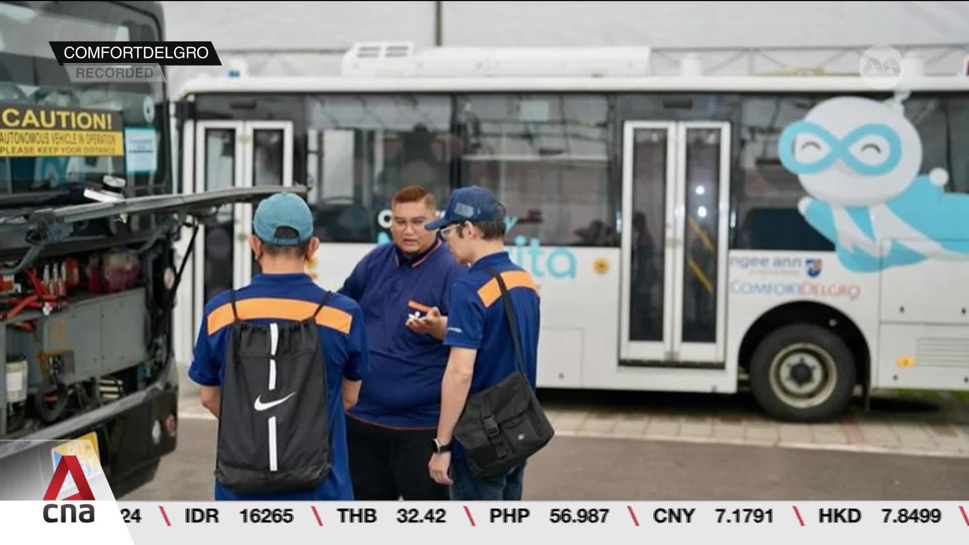 Three men in blue uniforms are gathered near an open bus engine compartment, discussing something.  A second bus, emblazoned with a cartoon figure, is parked nearby.
