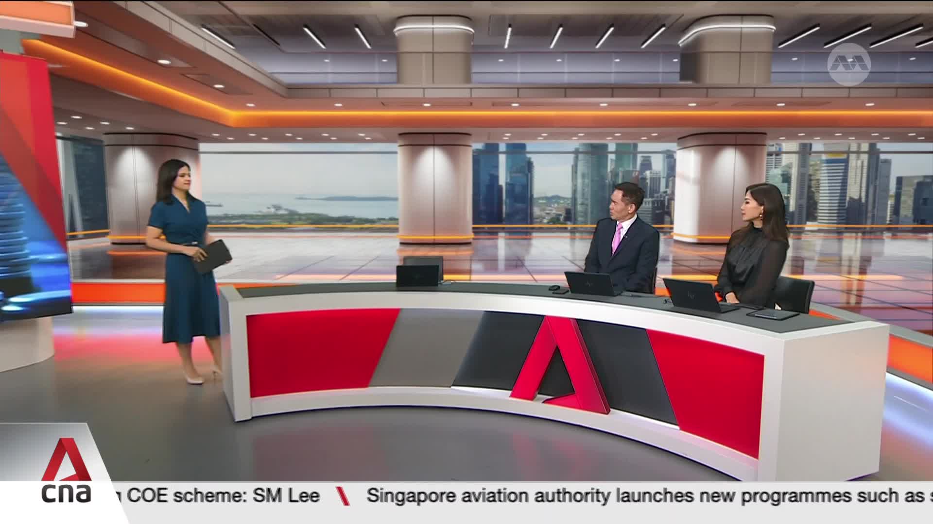 Three people are seated at a curved desk, each with a laptop in front of them, while another woman stands nearby holding a tablet. The backdrop reveals a cityscape, likely Singapore, visible through large windows.
