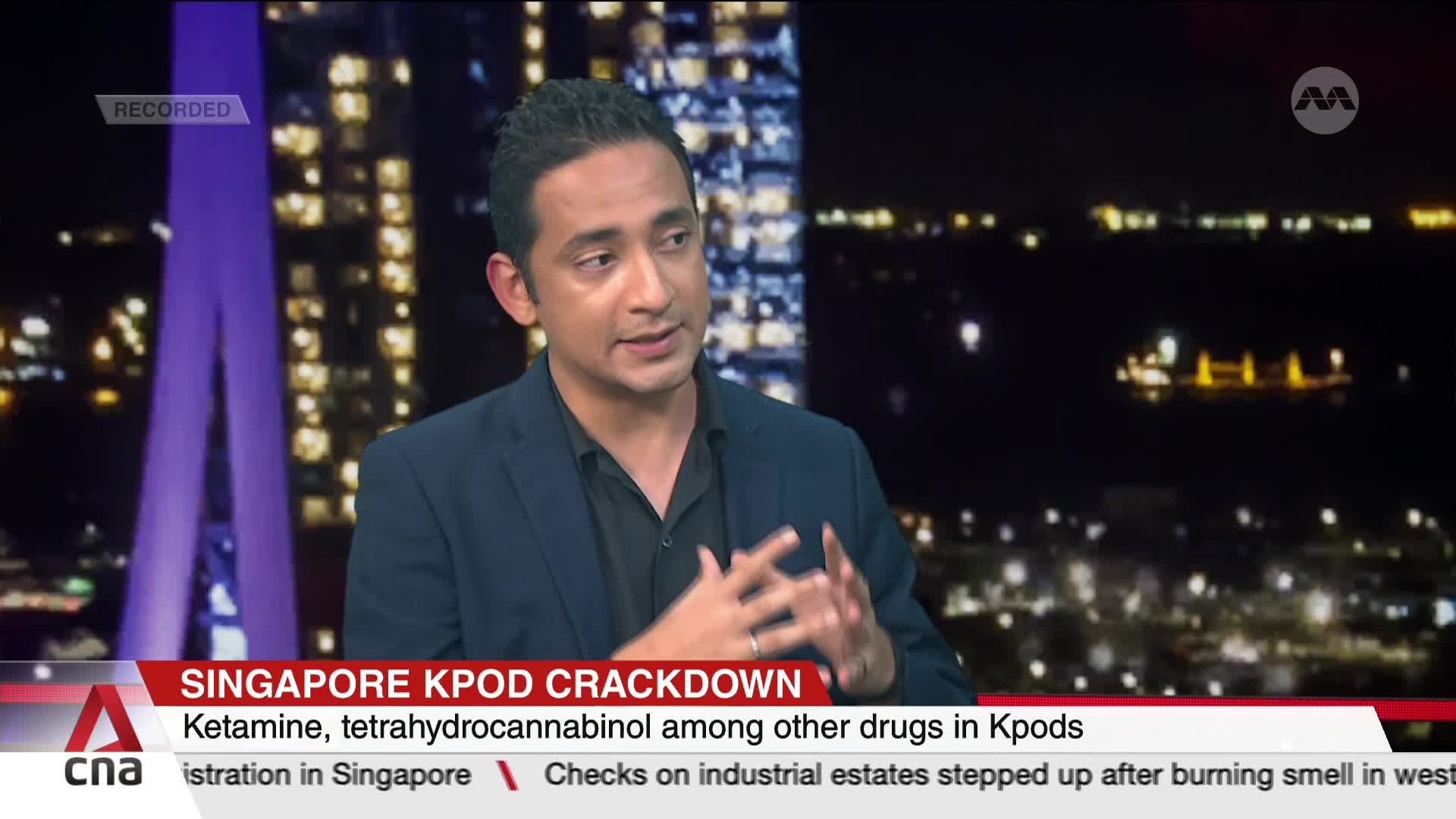 A man in a dark blazer gestures with his hands, speaking on a news segment from CNA International. The broadcast's lower third discusses a Singapore Kpod crackdown, with a backdrop of city lights.
