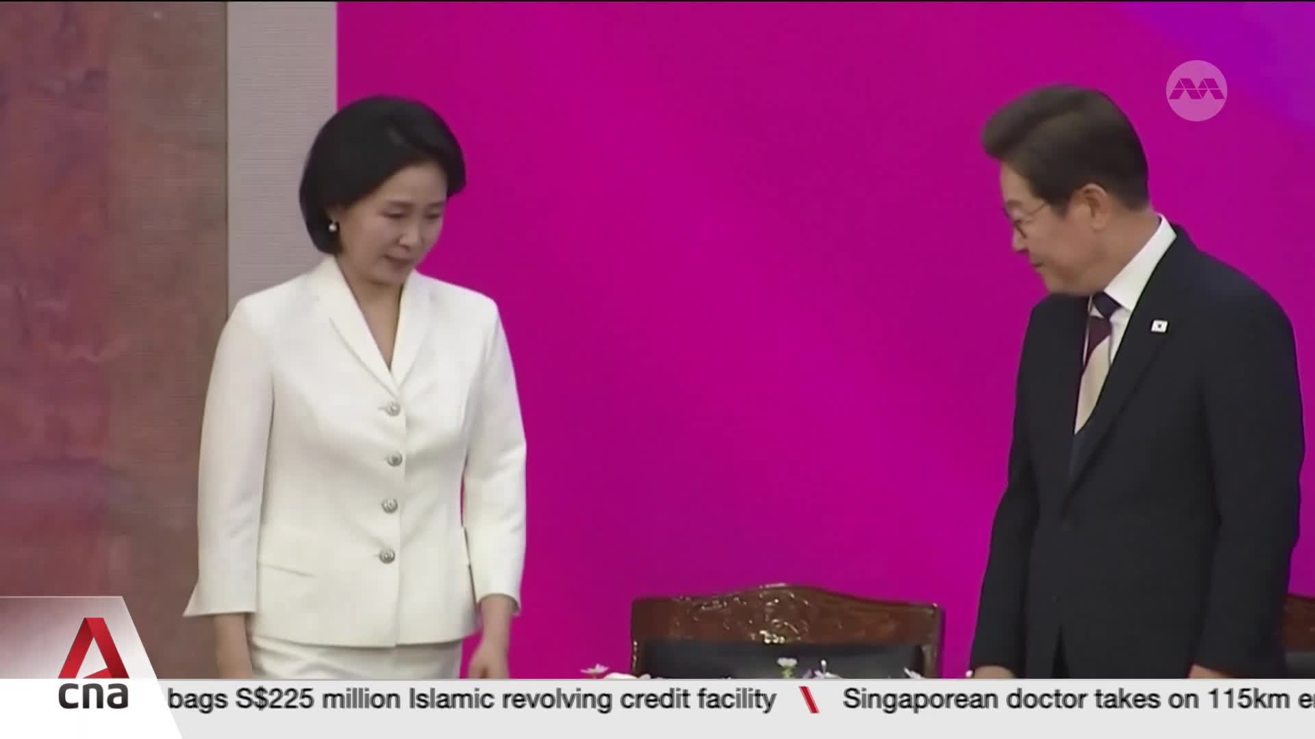 A woman in a white suit stands at the edge of a table, looking down. A man in a dark suit and glasses stands beside her, also looking down, as CNA International reports from Singapore.
