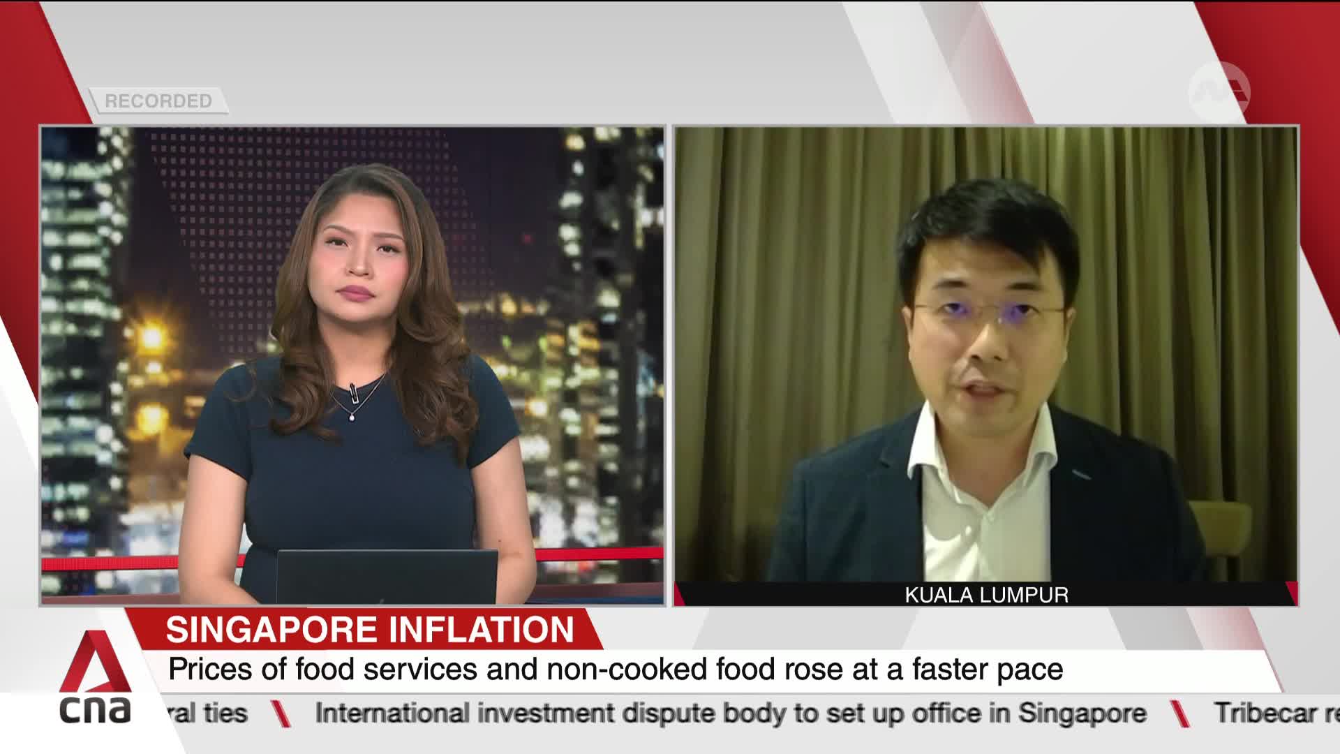 A news anchor in Singapore, on CNA International, is listening intently as a man in Kuala Lumpur speaks, likely about Singapore's inflation. The lower third of the screen displays a headline about rising food prices.
