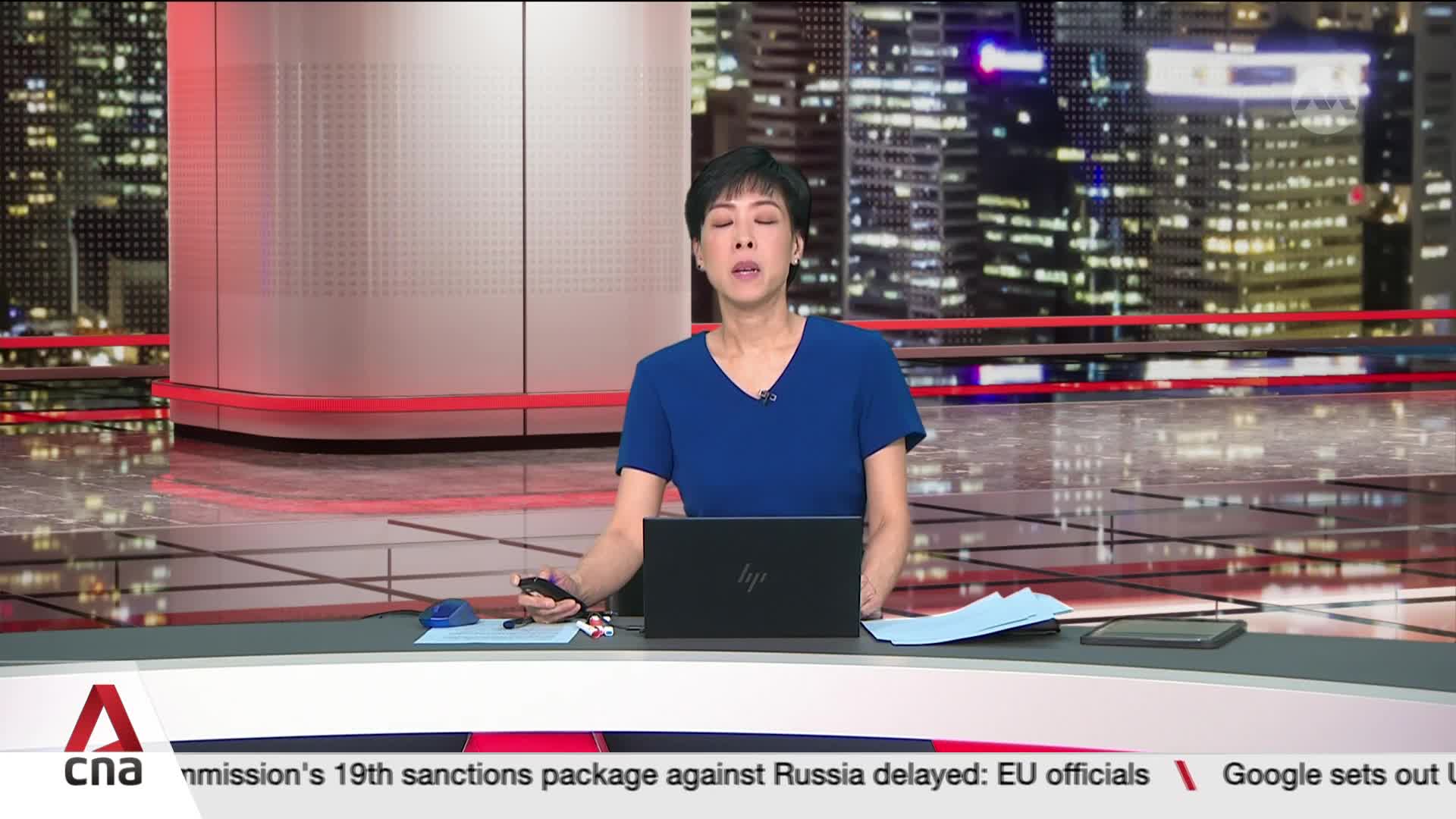 A woman in a blue top speaks directly to the camera, a laptop and papers arranged on the desk in front of her. The CNA logo is visible in the lower left corner, with news headlines scrolling beneath.
