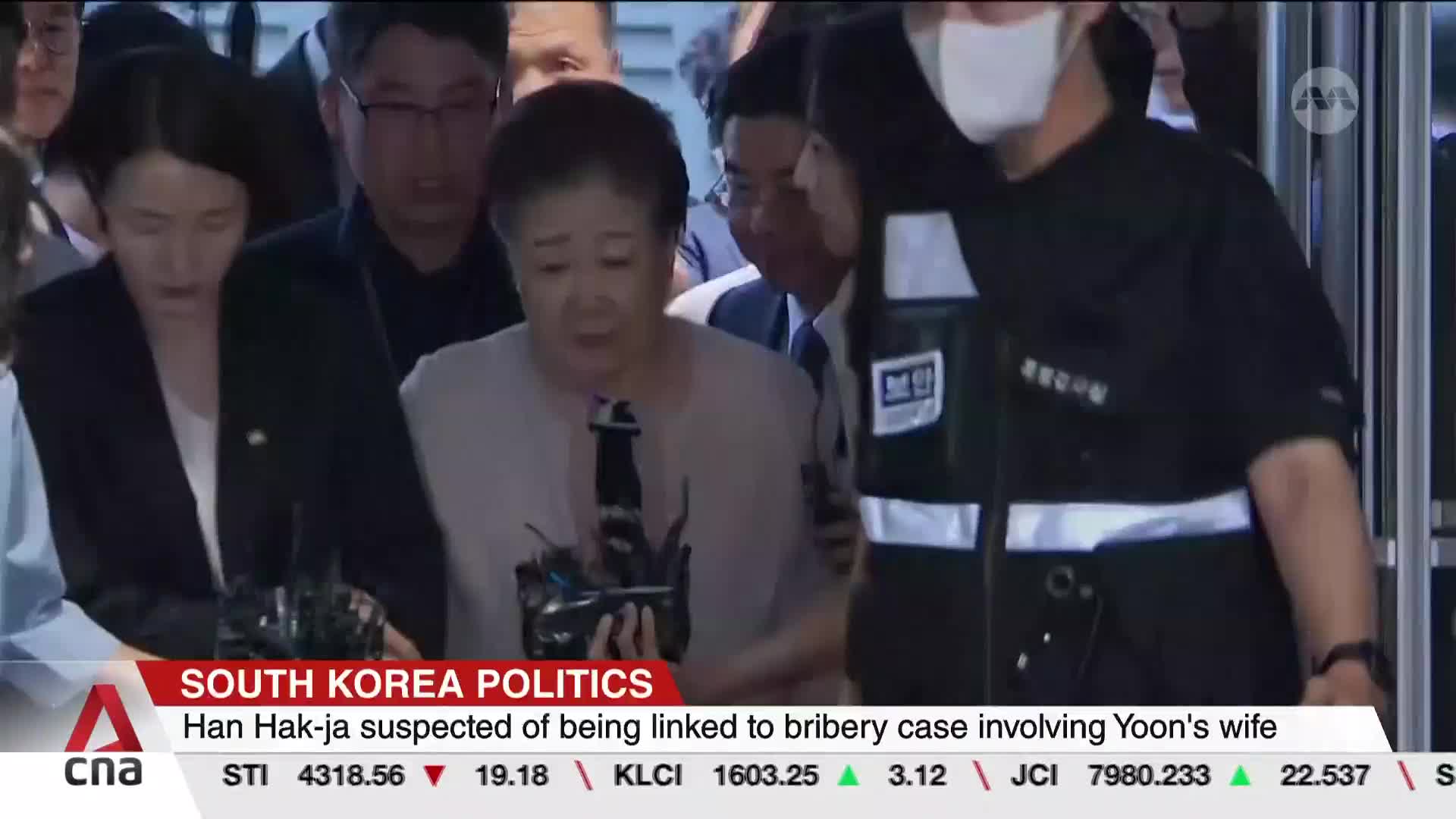 Amidst a throng of people, a woman is being escorted, her face etched with concern. The CNA International news ticker at the bottom provides context for the scene.
