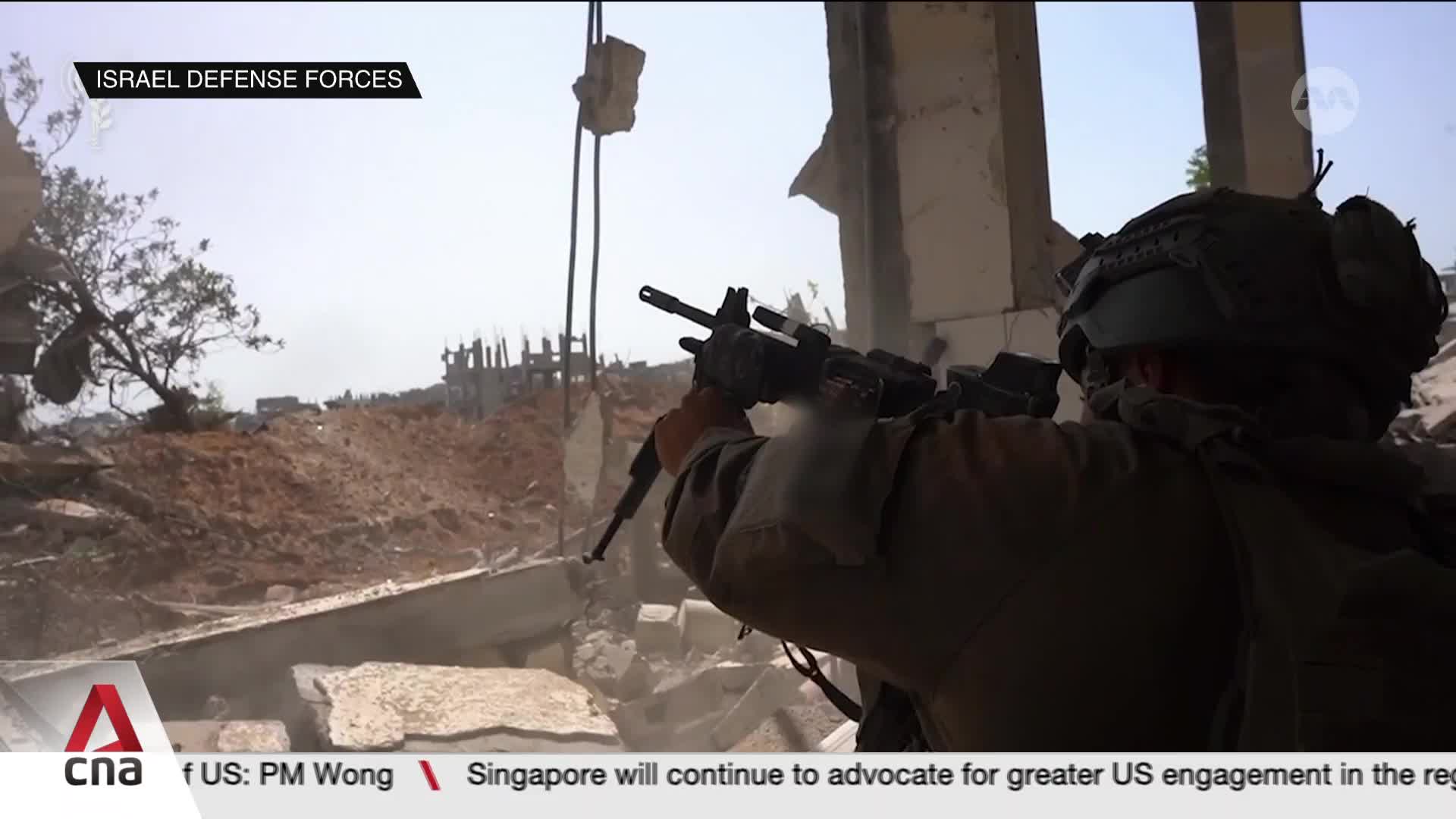 An Israeli soldier, seen from behind, aims his rifle through a broken structure. Debris litters the ground, and the scene is broadcast by CNA International.
