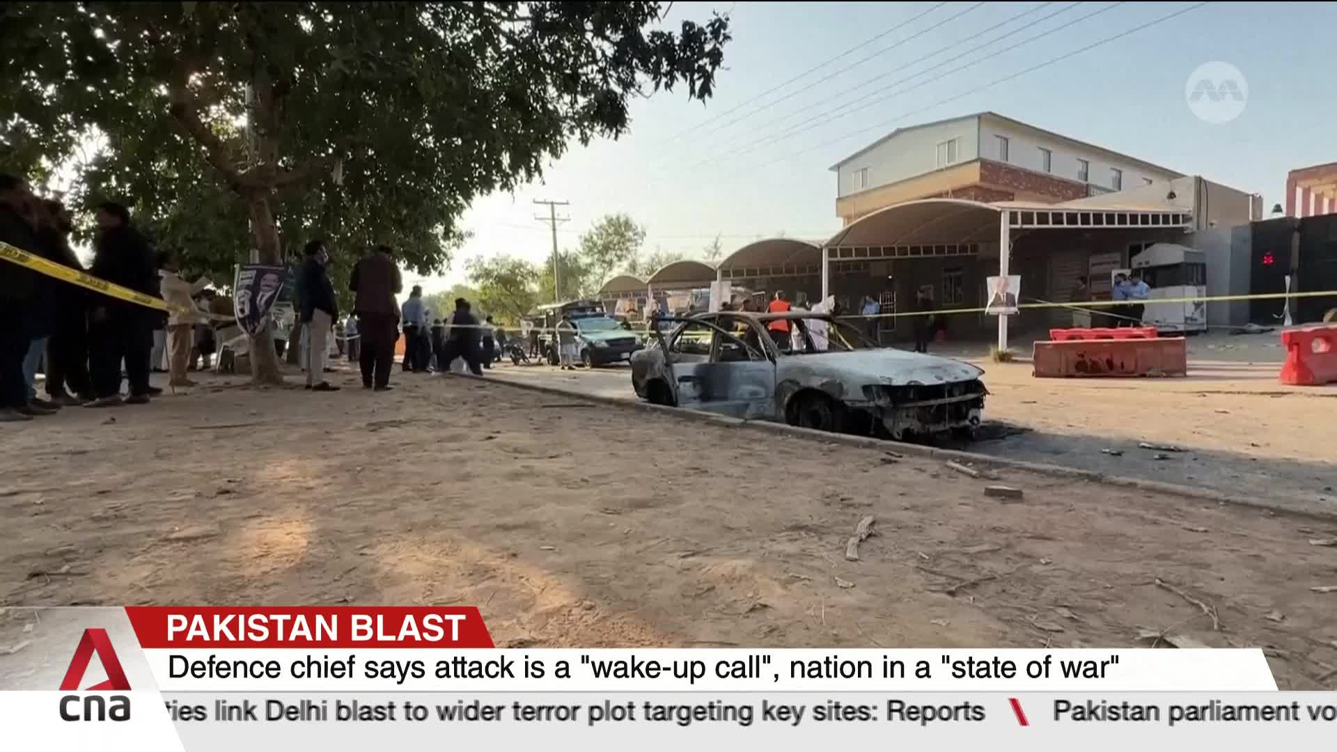 A burnt-out car sits on the side of the road, cordoned off with yellow tape. People stand and mill around, some looking towards the scene, as CNA International reports on the attack.
