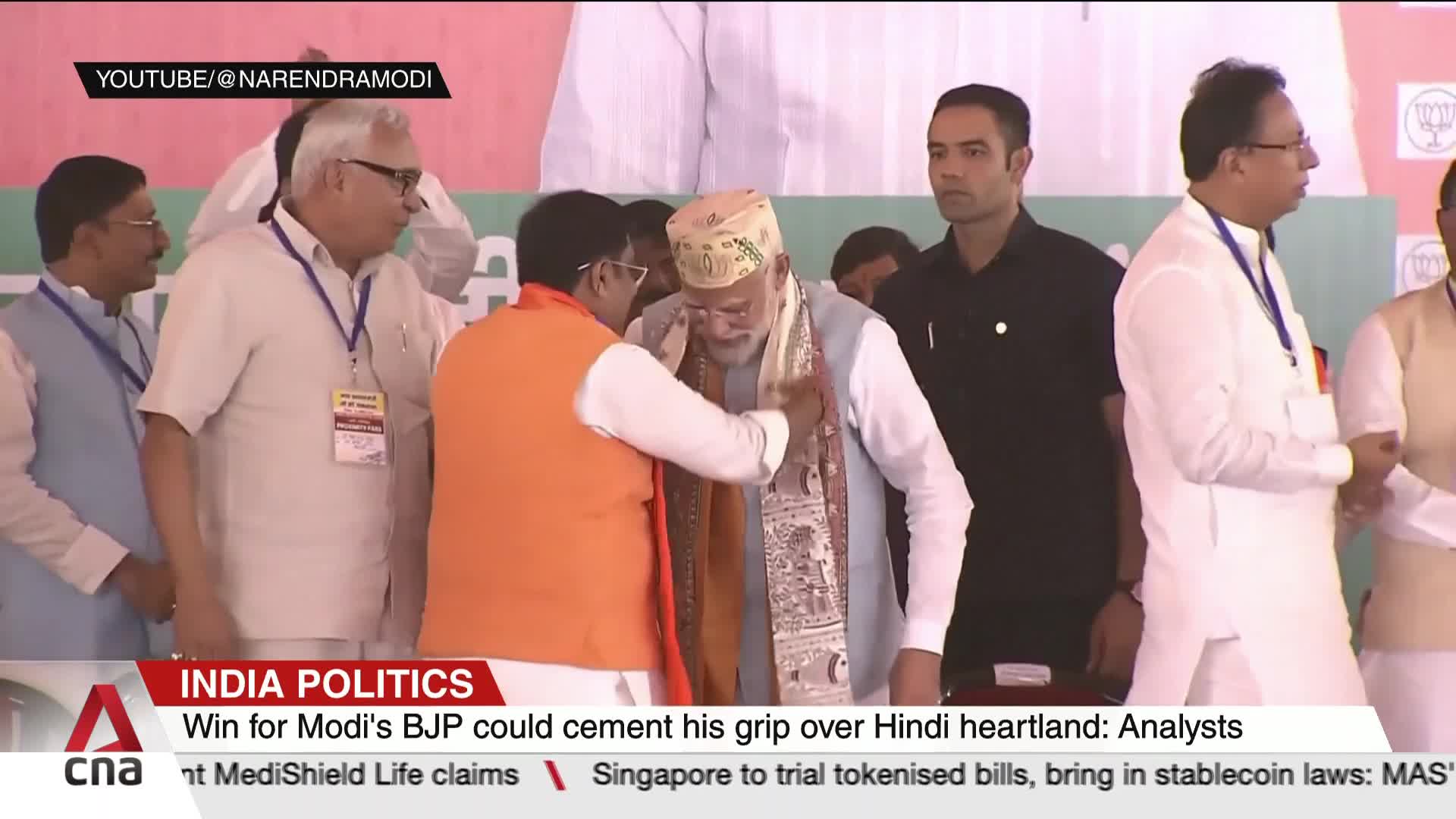 A man in an orange vest is draping a scarf around another man's neck. The man receiving the scarf is wearing a patterned hat and a light blue vest.
