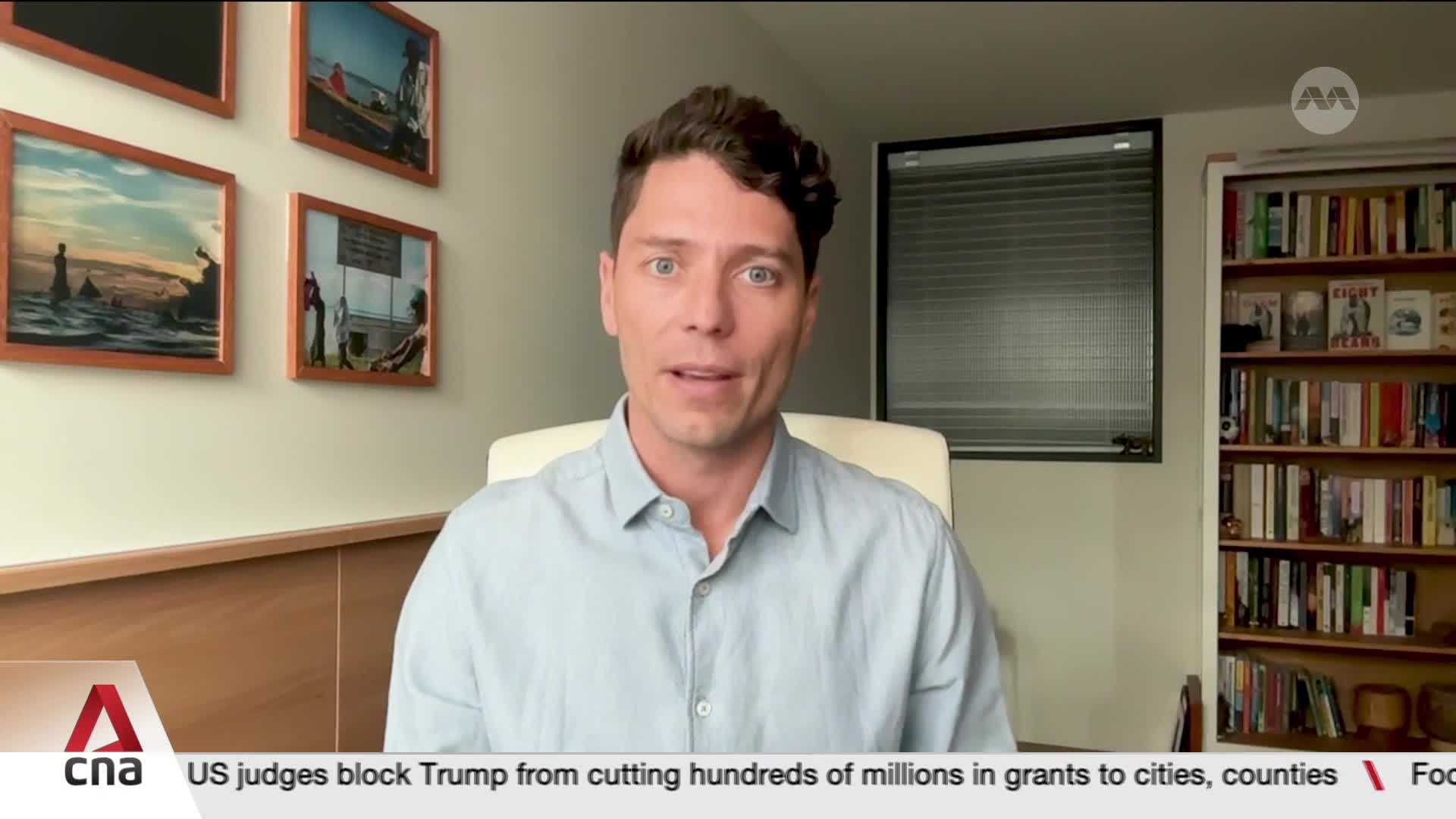 A man in a light blue shirt is speaking directly to the camera, likely for a CNA International broadcast. Behind him, a bookshelf and framed photos add context to the scene.
