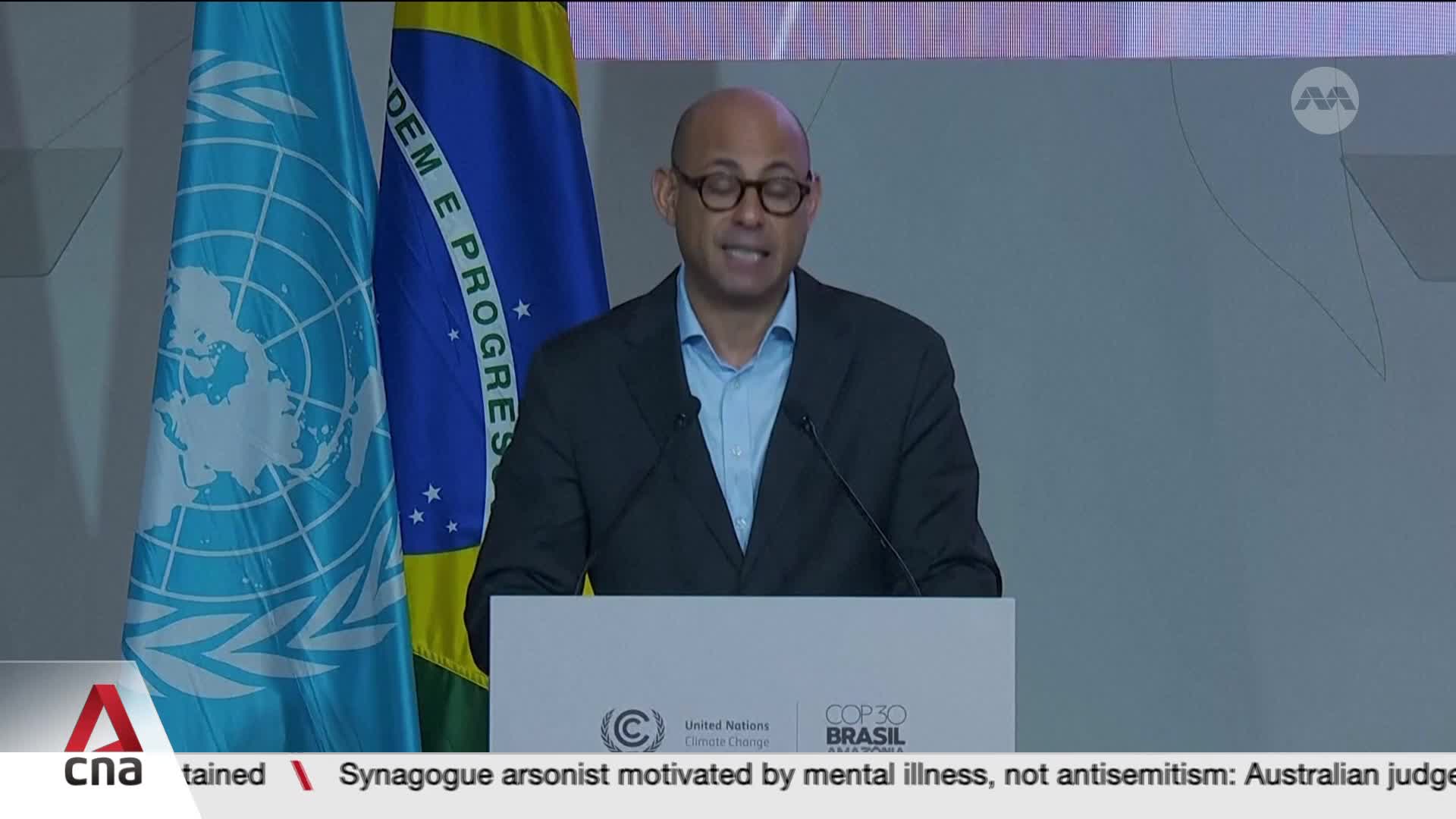 A man in glasses and a dark suit stands at a podium, speaking. Behind him, the flags of the United Nations and Brazil are displayed, and the CNA International logo is visible in the corner.
