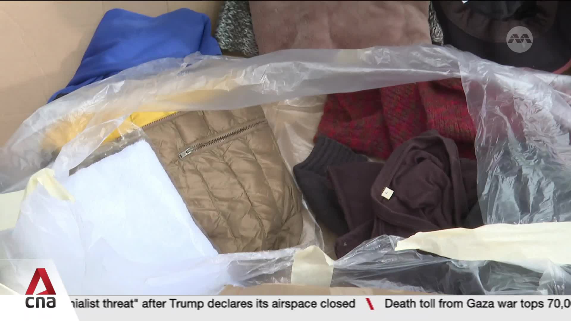 Inside a clear plastic bag, clothes are piled together, including a blue sweatshirt, a tan quilted vest, and a red patterned sweater. The CNA International logo is visible in the lower left corner.
