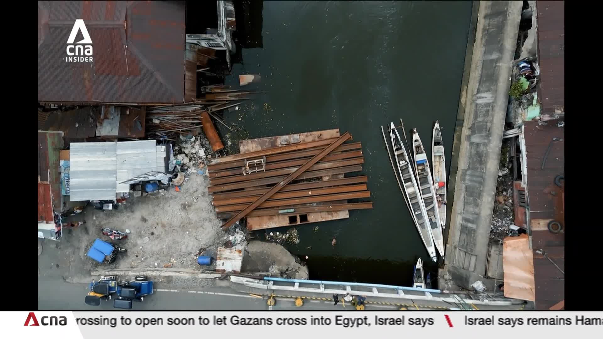 Rust-colored metal beams are stacked on a small platform near the water's edge. Three long, slender boats are moored alongside a concrete structure, seemingly ready to launch.
