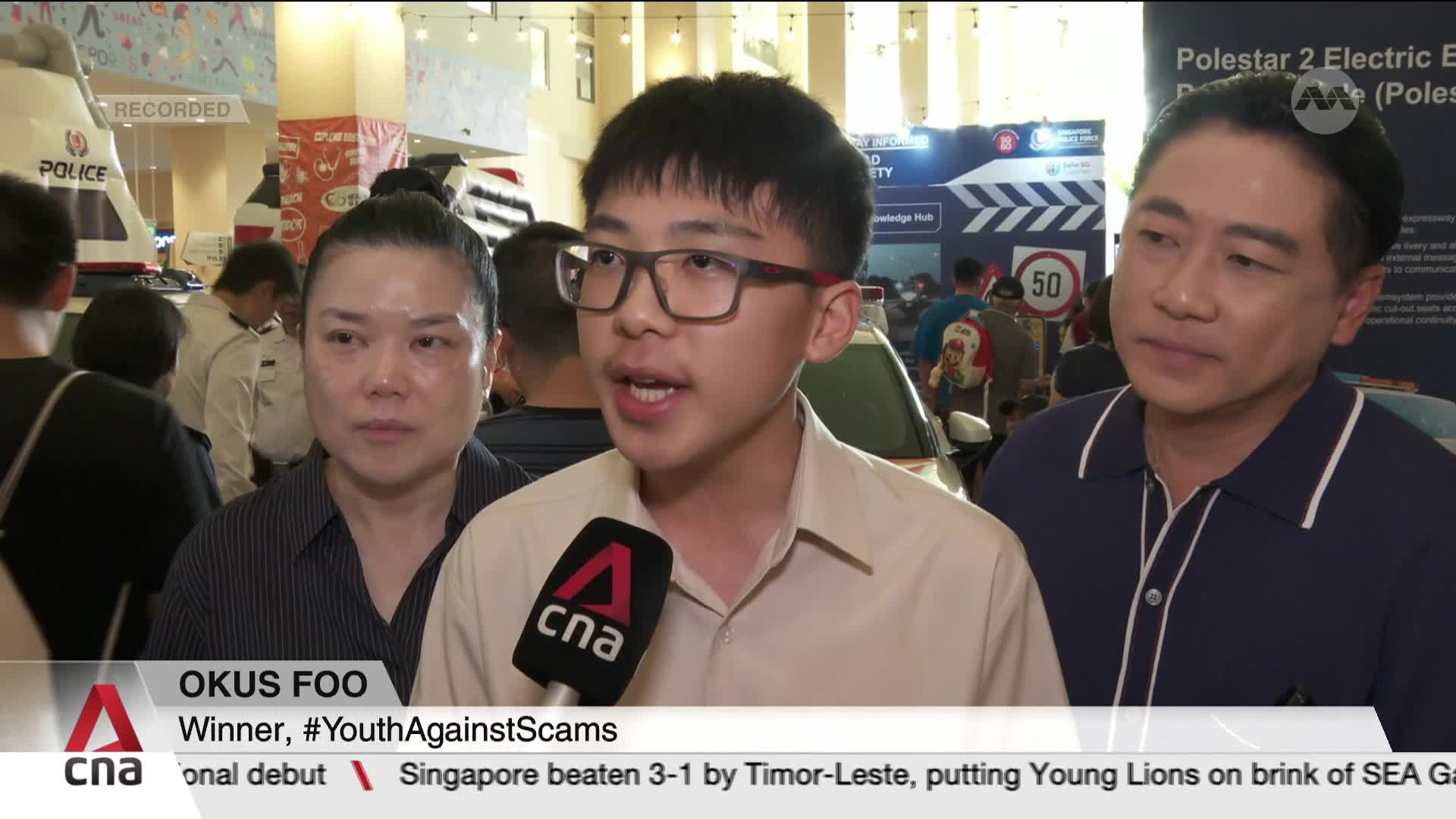 A young man, wearing glasses, speaks into a CNA microphone, flanked by two others. Behind them, a police vehicle display and a sign about electric vehicles are visible.
