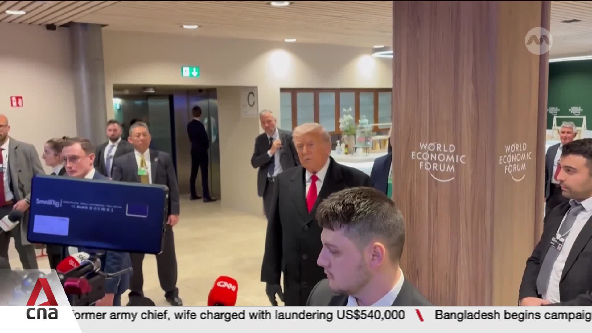 Donald Trump, in a black coat and red tie, walks past a "World Economic Forum" pillar. A group of reporters, with CNA International branding visible, are positioned to his left, holding microphones and a camera.