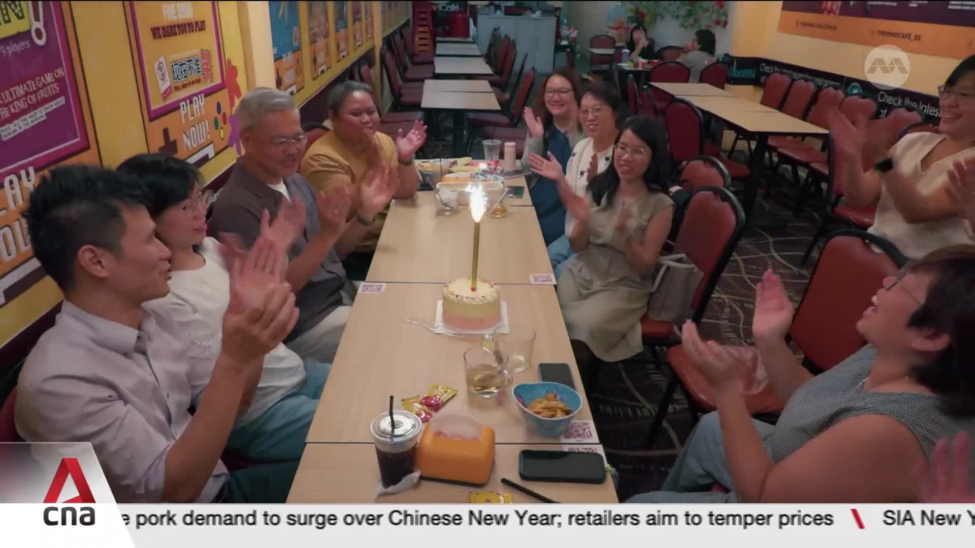 A small cake with a single lit candle sits on the table as everyone around it claps. The group, gathered in a Singapore restaurant, smiles and applauds the celebration. A small cake with a single lit candle sits on the table as everyone around it claps. The group, gathered in a Singapore restaurant, smiles and applauds the celebration.