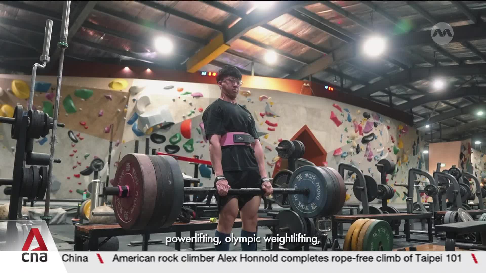 A young man grips a loaded barbell, his back straight and his face determined as he prepares for a deadlift. The gym walls behind him are covered in colorful climbing holds, a stark contrast to the heavy weights he's about to lift. A young man grips a loaded barbell, his back straight and his face determined as he prepares for a deadlift. The gym walls behind him are covered in colorful climbing holds, a stark contrast to the heavy weights he's about to lift.
