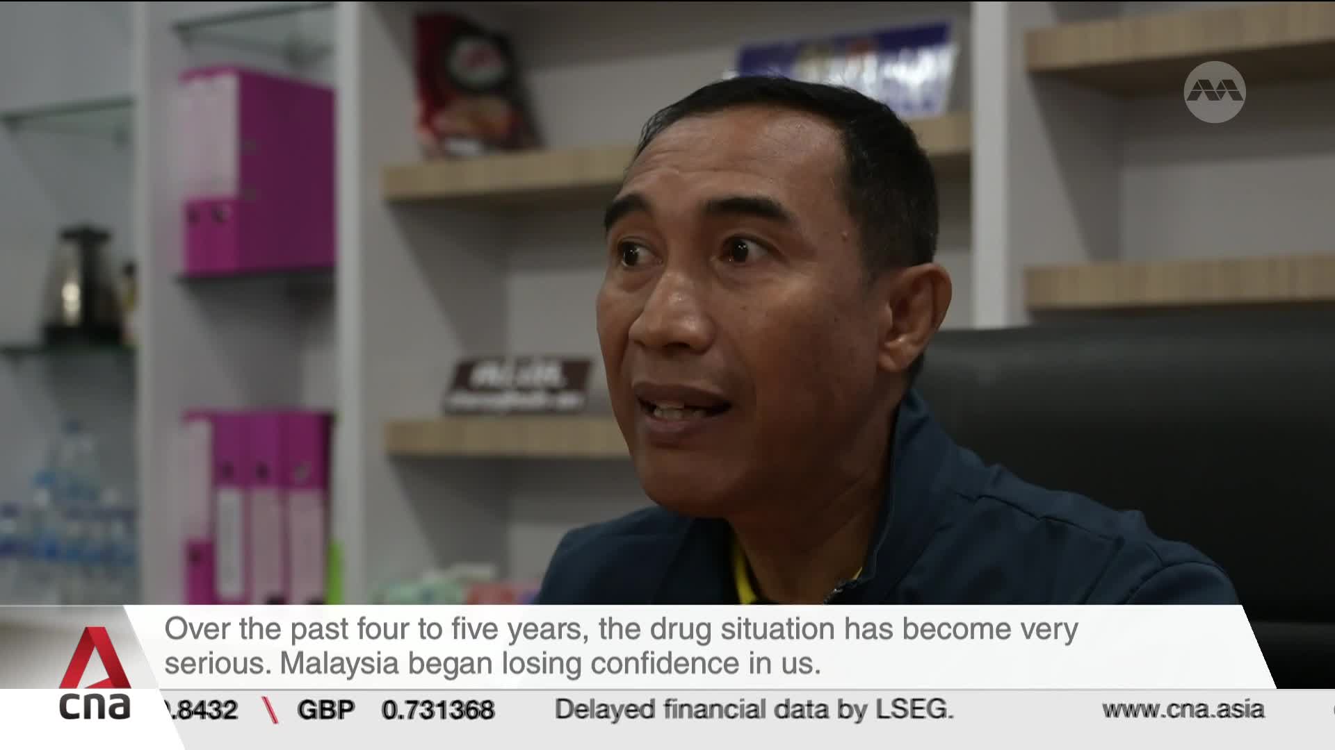 A man speaks directly to the camera, his expression earnest. Behind him, shelves hold binders and what looks like a coffee maker. A man speaks directly to the camera, his expression earnest. Behind him, shelves hold binders and what looks like a coffee maker.