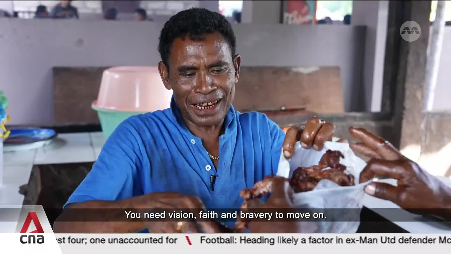 A man in a blue shirt smiles as he holds out a piece of food wrapped in white paper. Another hand reaches in to take it. A man in a blue shirt smiles as he holds out a piece of food wrapped in white paper. Another hand reaches in to take it.