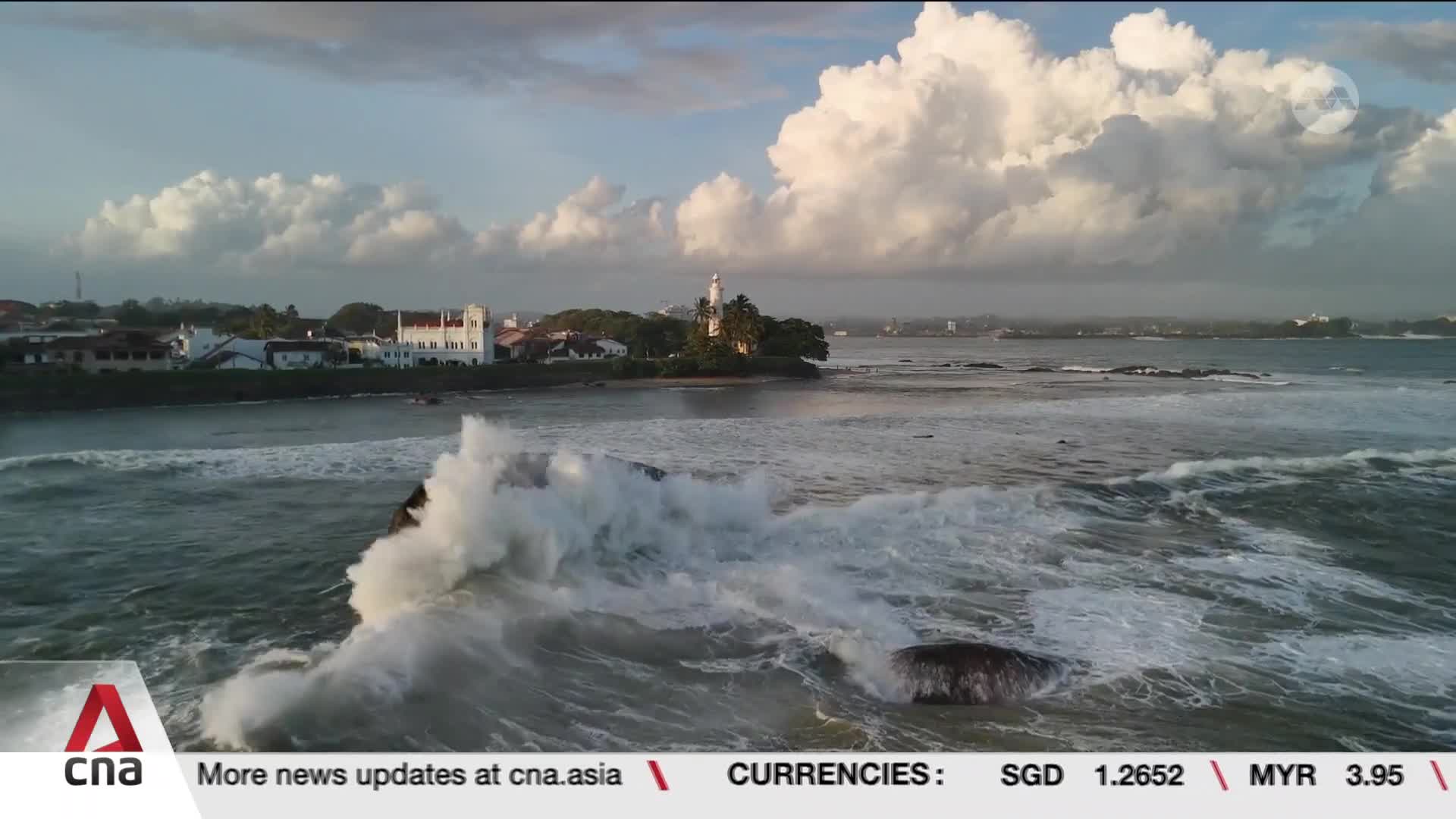 Waves crash against dark rocks, sending white spray high into the air. In the distance, a white lighthouse stands against a backdrop of colonial-era buildings. Waves crash against dark rocks, sending white spray high into the air. In the distance, a white lighthouse stands against a backdrop of colonial-era buildings.