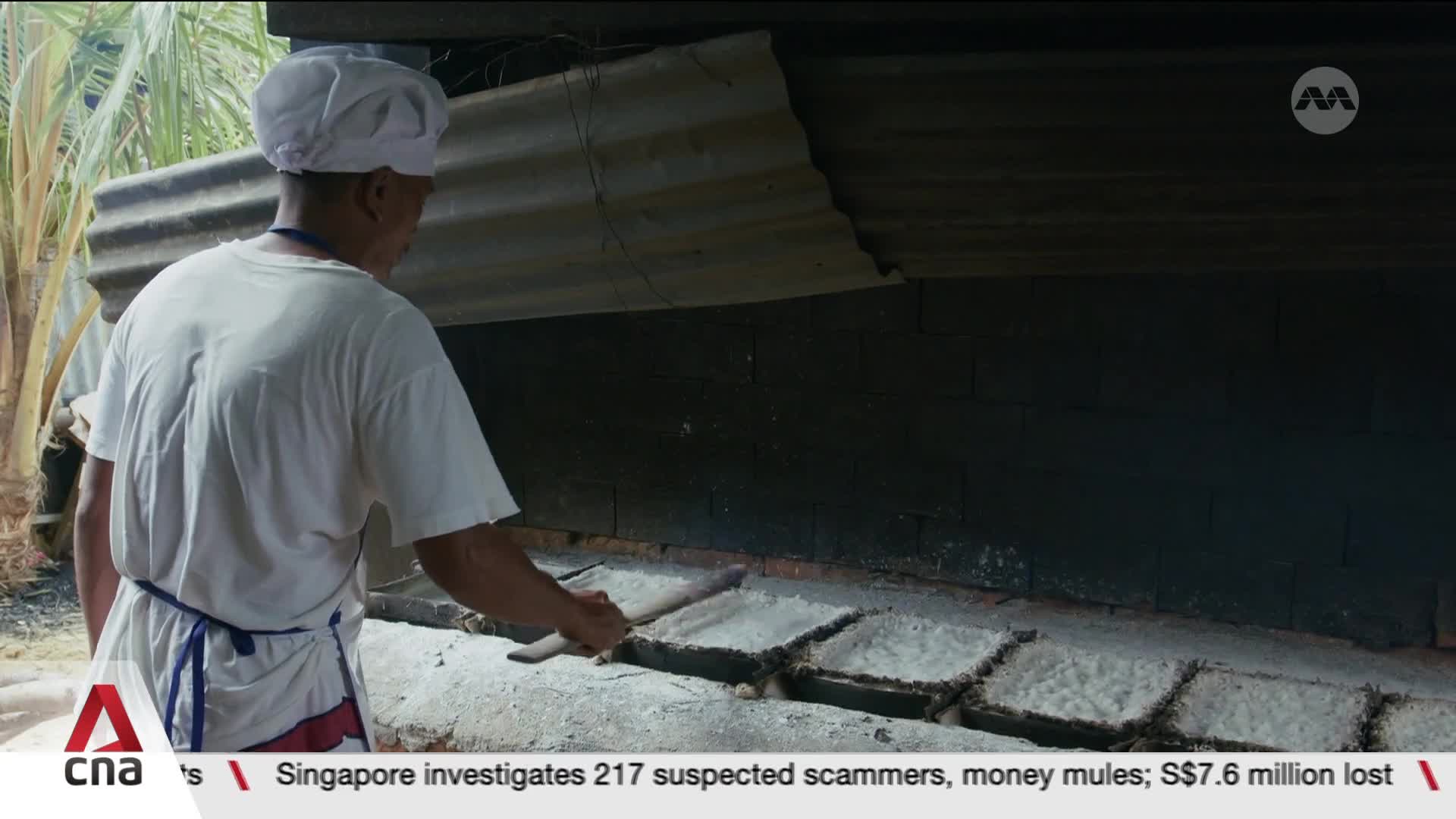 A man in a white chef's hat and apron carefully arranges square trays of what looks like salt onto a heated surface. The trays are laid out in a row, ready for processing in this Singaporean setting. A man in a white chef's hat and apron carefully arranges square trays of what looks like salt onto a heated surface. The trays are laid out in a row, ready for processing in this Singaporean setting.