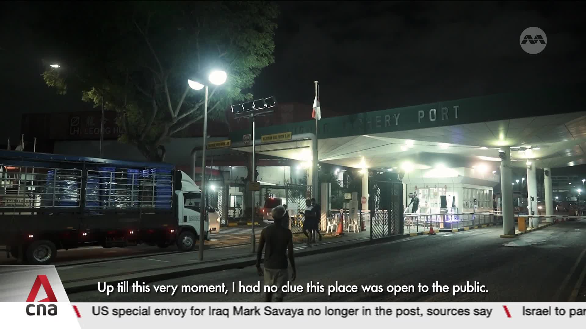 A large truck with a caged back is parked near the entrance to a port facility. People stand around the brightly lit entrance, some looking towards the road. A large truck with a caged back is parked near the entrance to a port facility. People stand around the brightly lit entrance, some looking towards the road.