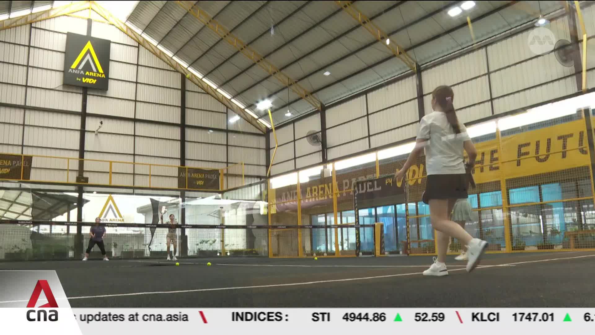 Two people are playing padel on a court in Singapore. The woman in the foreground is about to swing at a ball. Two people are playing padel on a court in Singapore. The woman in the foreground is about to swing at a ball.