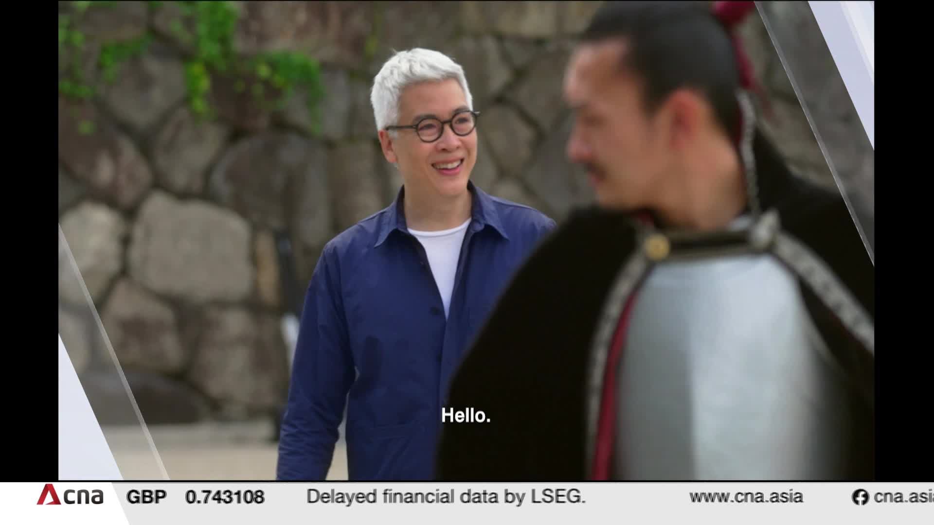 A man with bright white hair and glasses smiles, looking at someone in historical armor. The scene unfolds outdoors, with a stone wall and greenery behind them. A man with bright white hair and glasses smiles, looking at someone in historical armor. The scene unfolds outdoors, with a stone wall and greenery behind them.