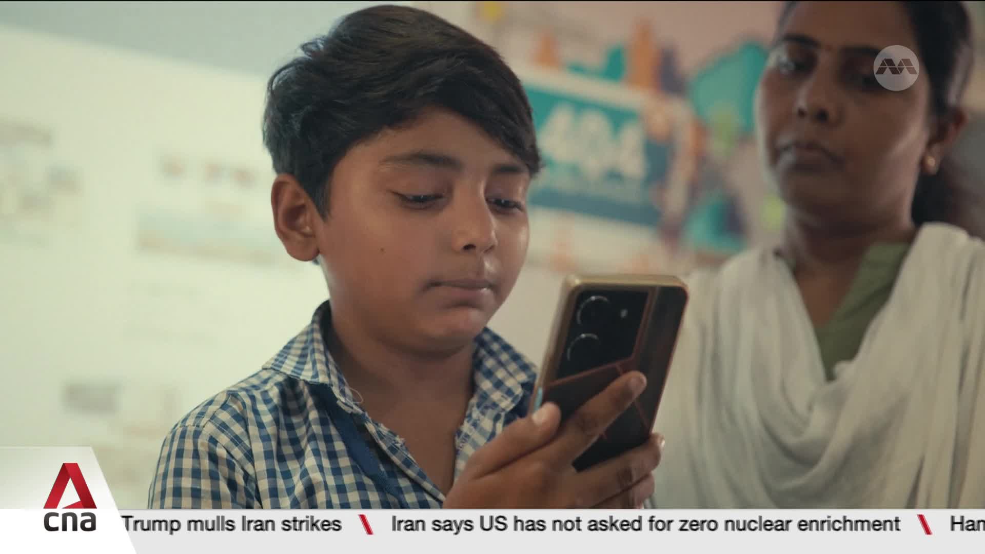 A young boy in Singapore, wearing a checkered shirt, intently focuses on a smartphone held in his hand. Behind him, a woman in a white shawl looks on, her expression unreadable. A young boy in Singapore, wearing a checkered shirt, intently focuses on a smartphone held in his hand. Behind him, a woman in a white shawl looks on, her expression unreadable.