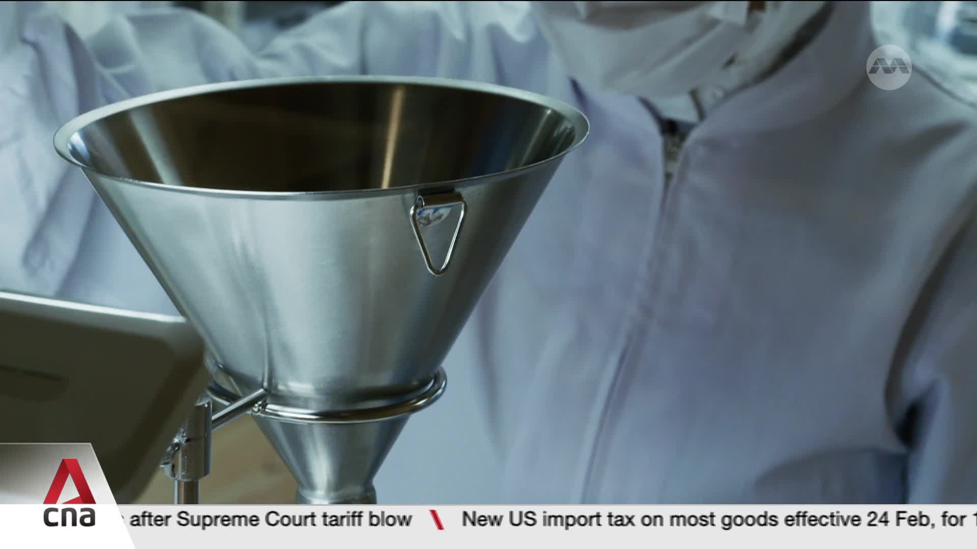 A person in a white lab coat is holding a large, shiny metal funnel. The CNA logo is visible in the bottom left corner. A person in a white lab coat is holding a large, shiny metal funnel. The CNA logo is visible in the bottom left corner.